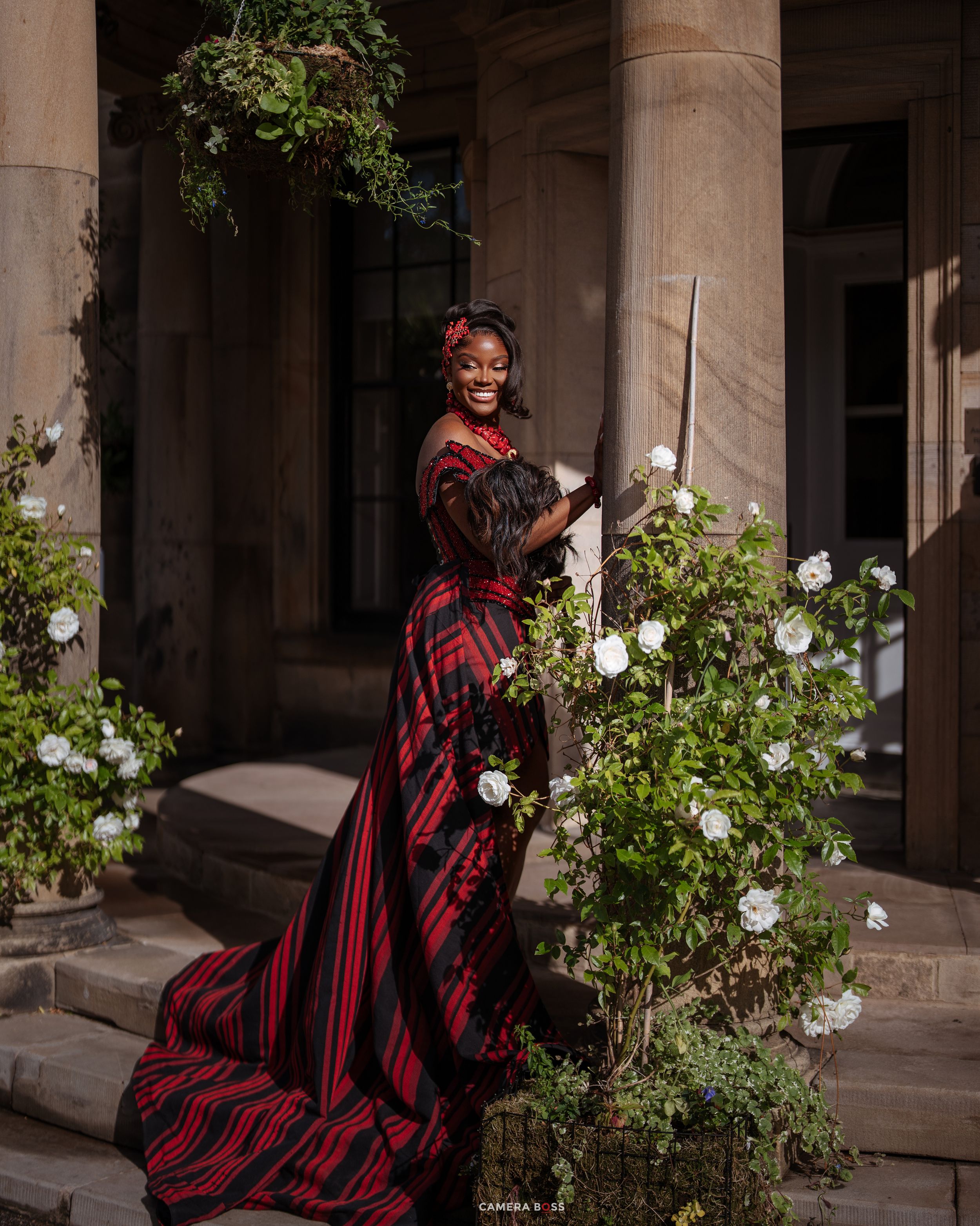Woman in red and black plaid gown smiling among white roses at elegant stone building.