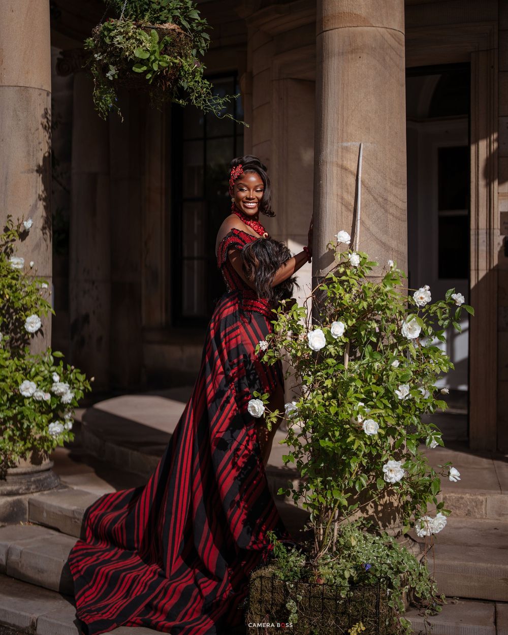 Woman in red and black plaid gown smiling among white roses at elegant stone building.