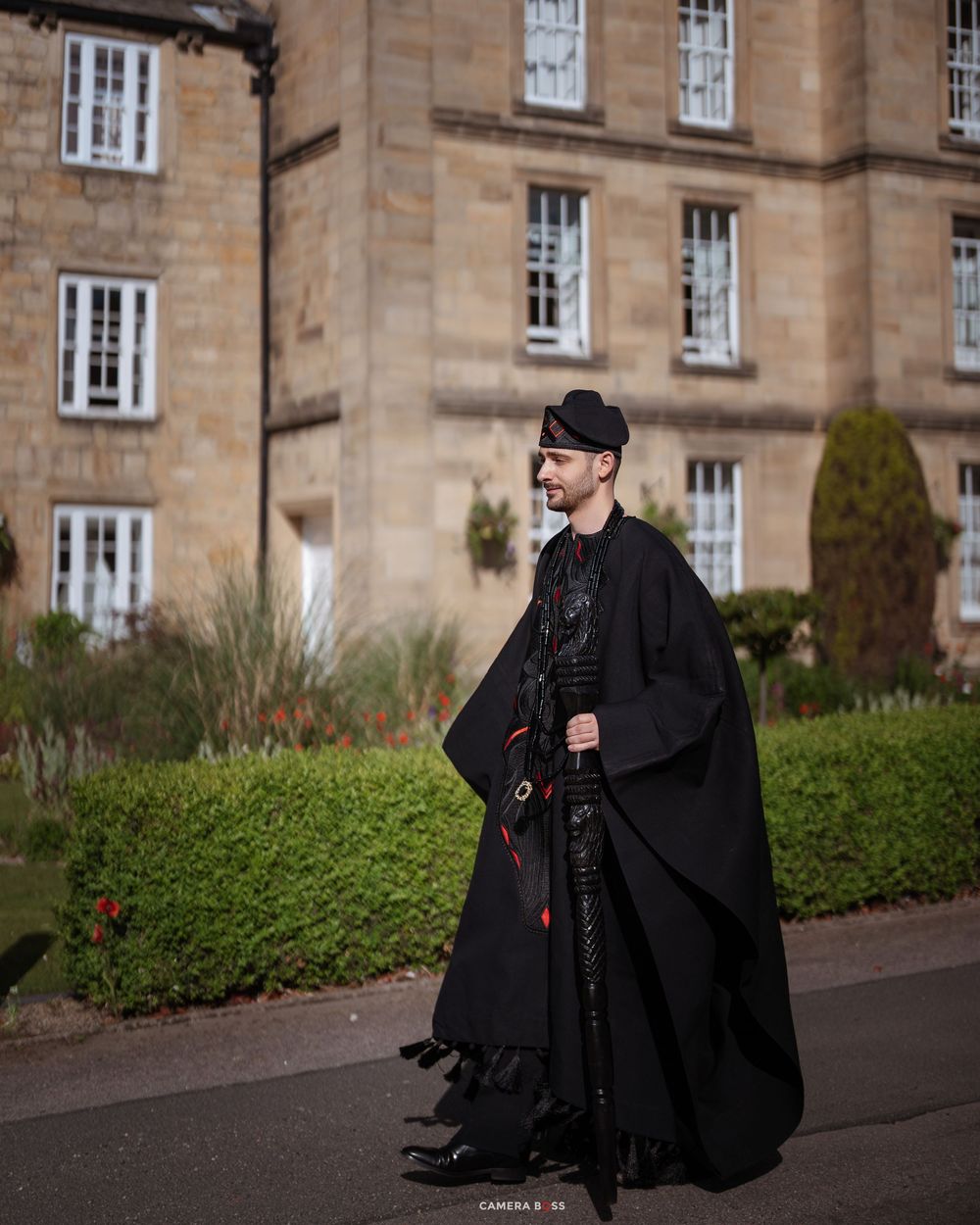 Man in elaborate black gothic cape and hat poses outside a historic stone building with manicured hedges.