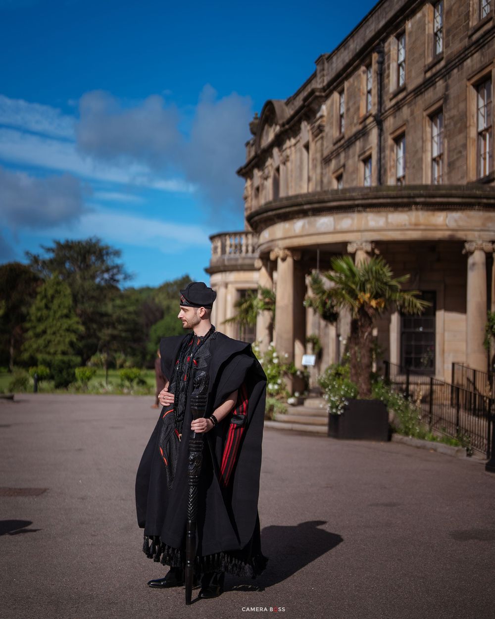 Man in black cape and flat cap poses before a grand stone building under blue skies.