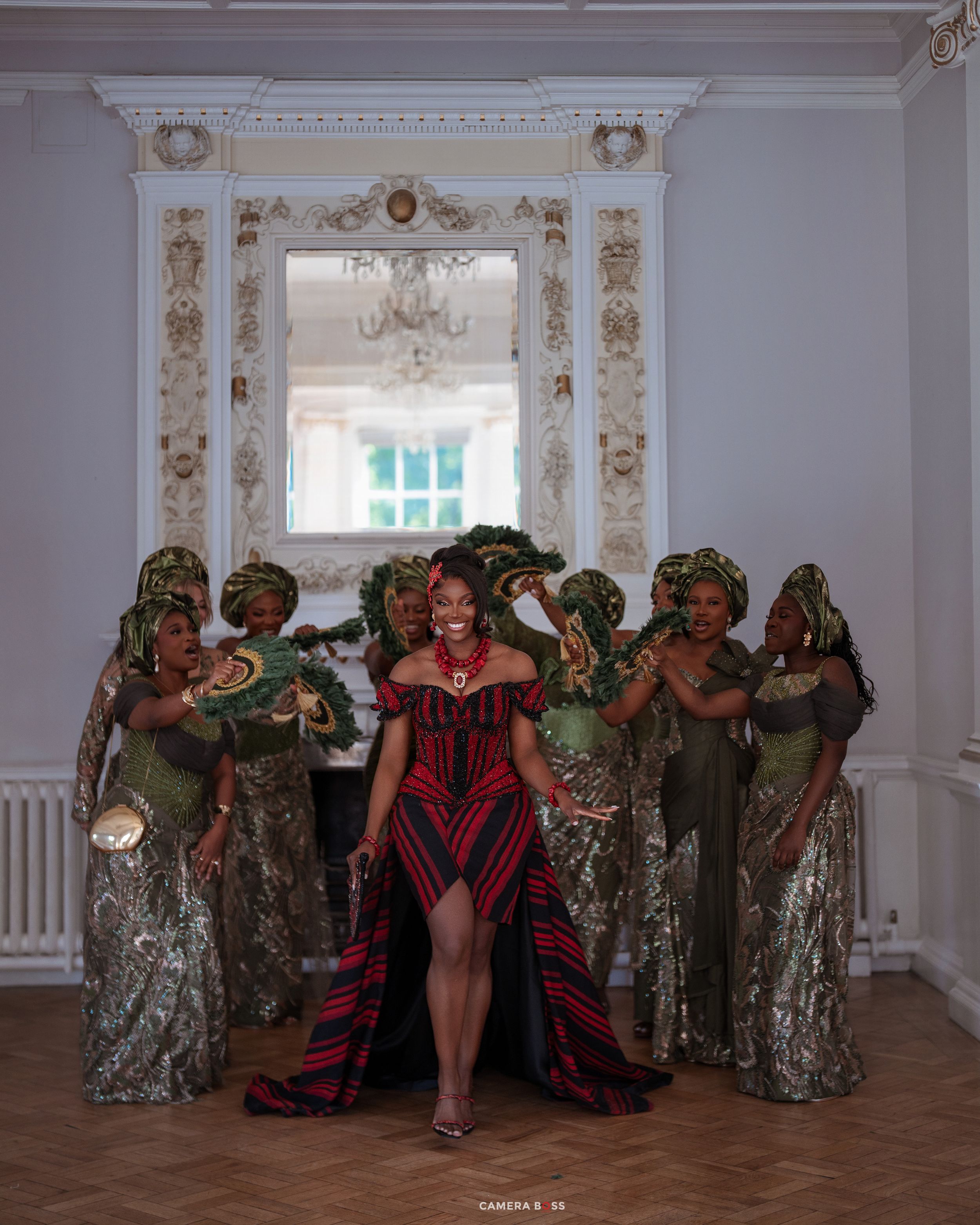 Bride in red traditional African dress with bridesmaids in green attire posing in elegant white room.