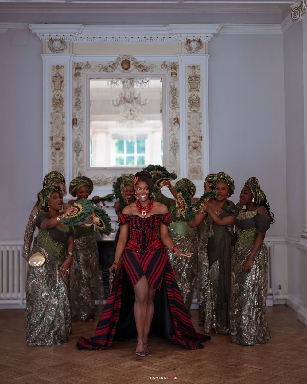 Bride in red traditional African dress with bridesmaids in green attire posing in elegant white room.