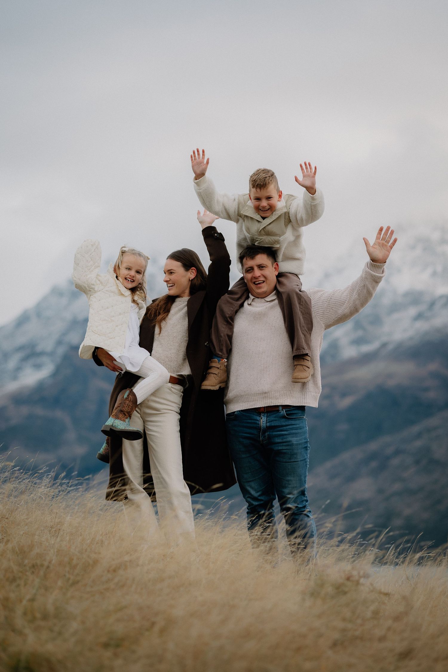 Happy family of four posing in a snowy mountain landscape, parents carrying children on their backs with arms raised.