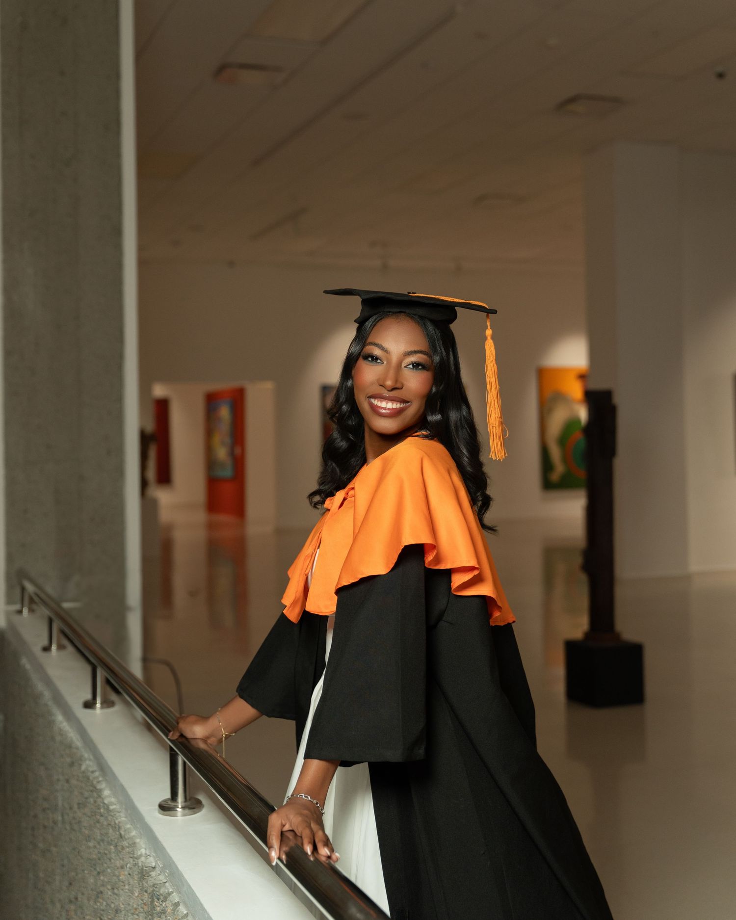 Smiling graduate in black cap and gown sits holding diploma folder, celebrating academic achievement.