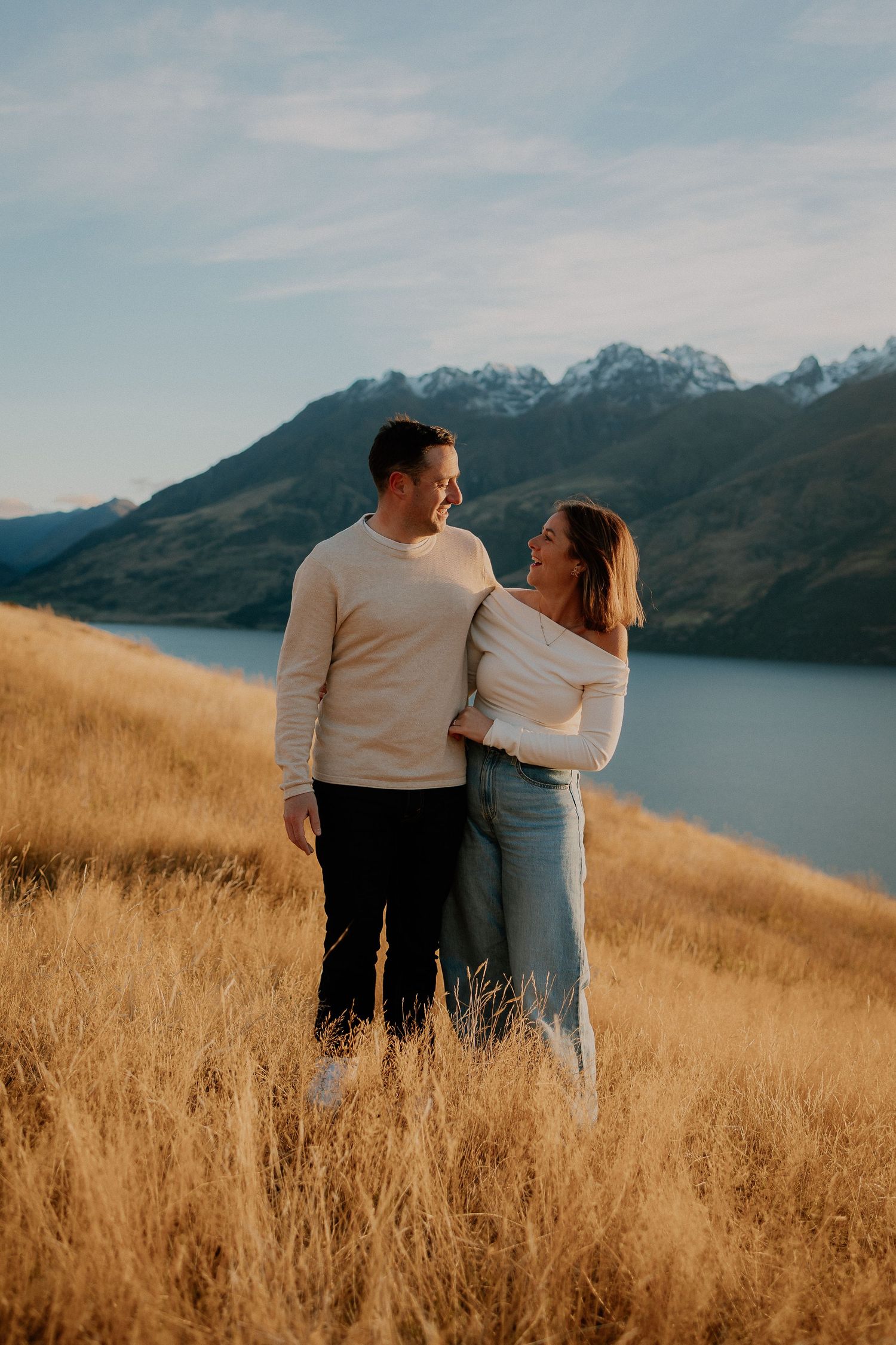 Couple embracing in golden grass field with snow-capped mountains and lake backdrop in New Zealand.