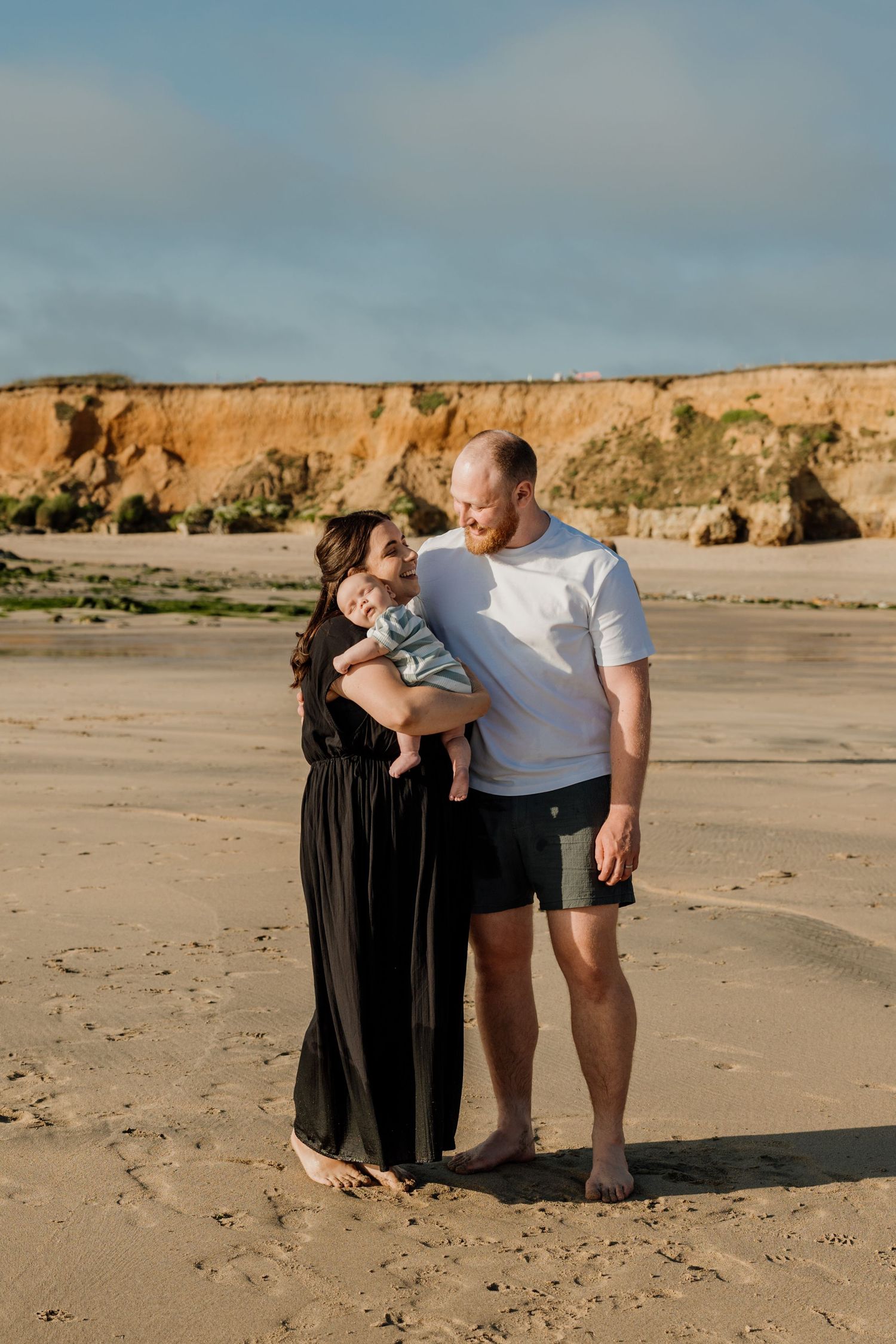 A young family with a baby poses on a sandy beach with golden cliffs in the background on a sunny day.
