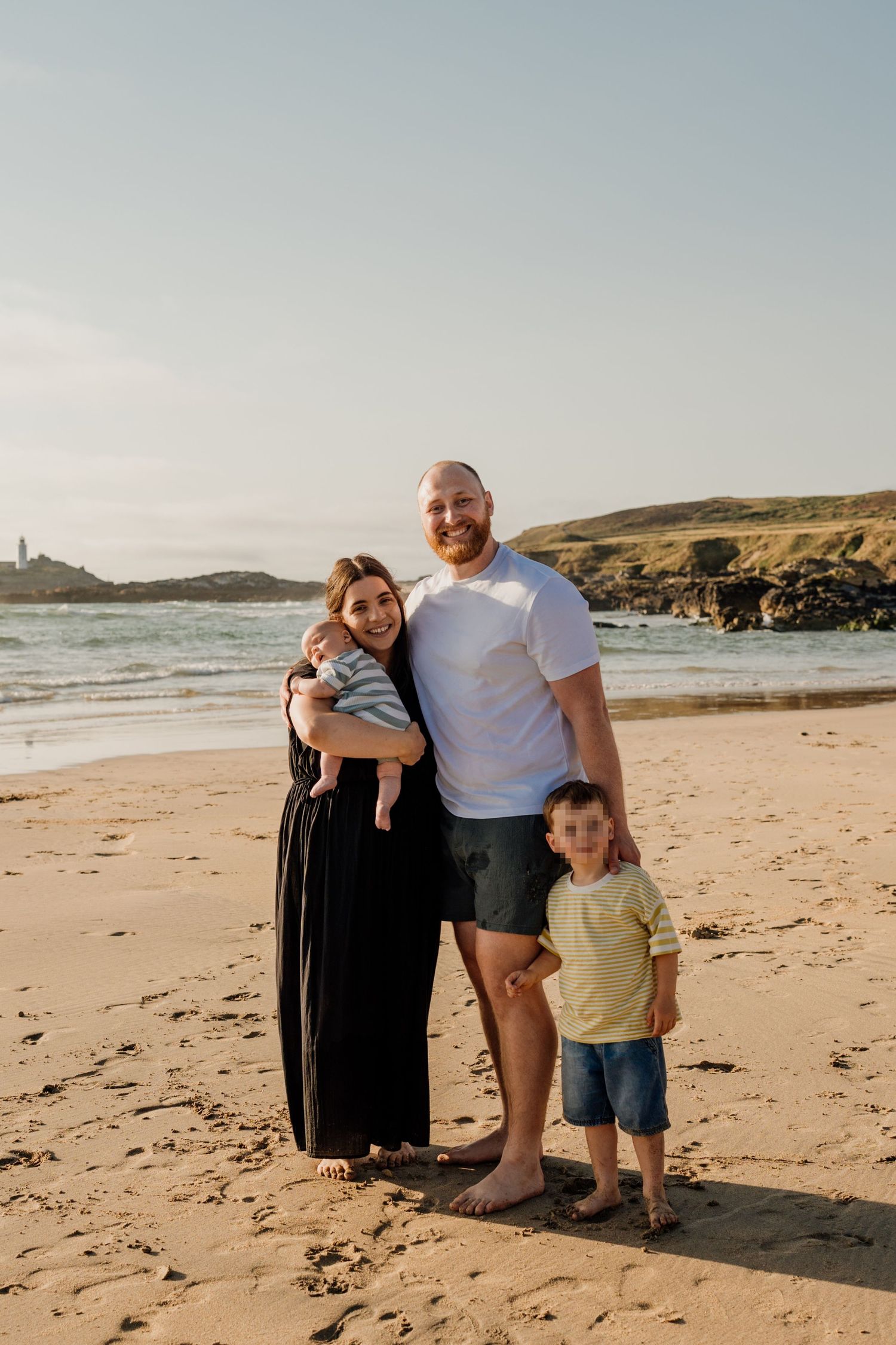 Family of four posing on sandy beach at golden hour, parents holding baby with young boy standing in front.