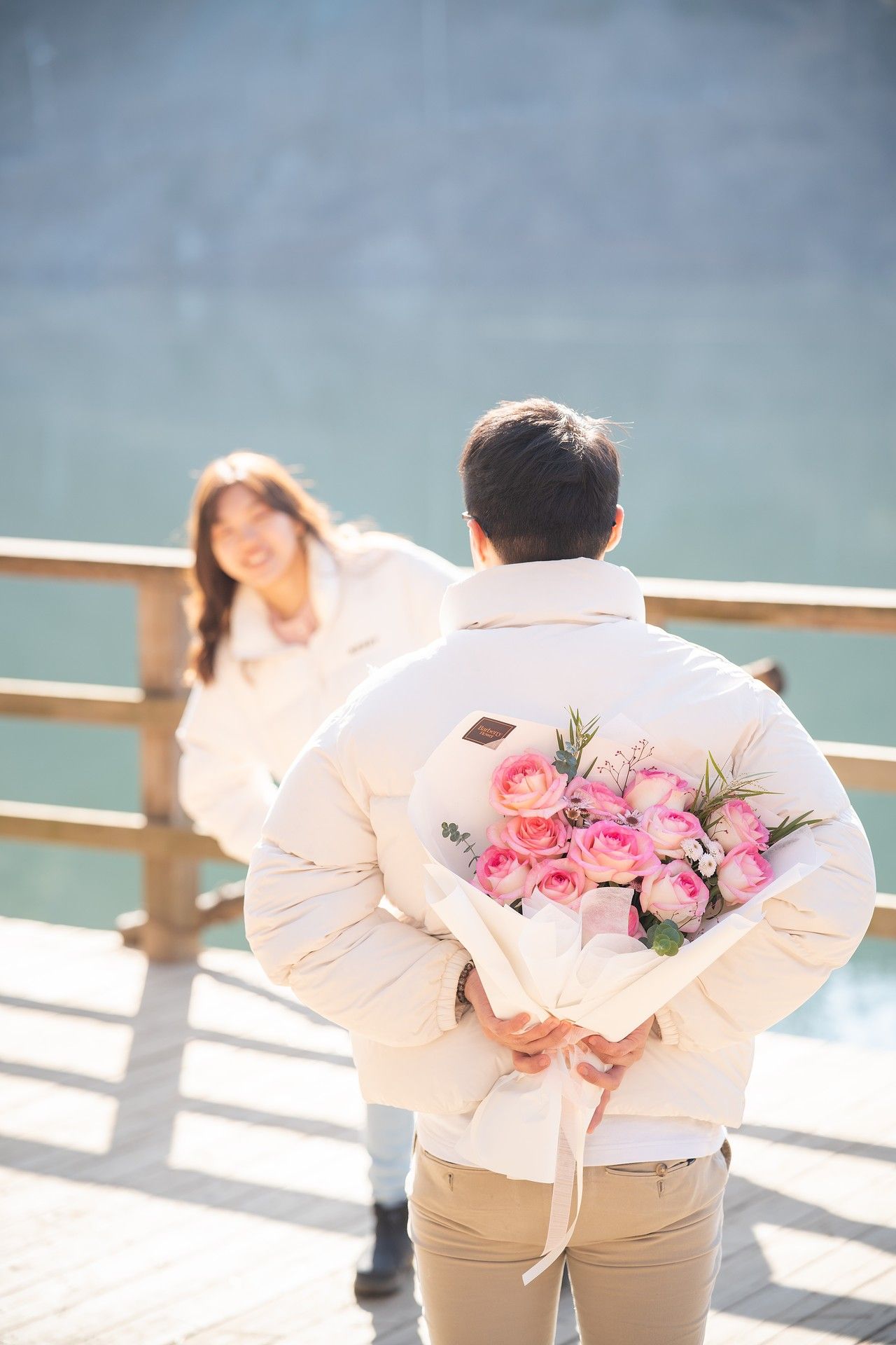 Man hiding pink rose bouquet behind his back to surprise a smiling woman on a sunny waterfront boardwalk.