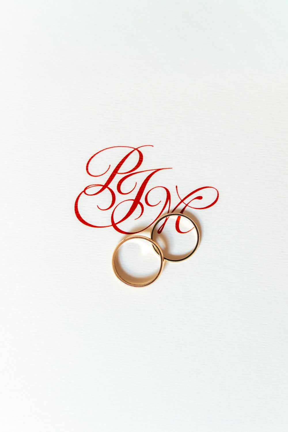 Two gold wedding bands beside red calligraphy initials on white textured background.