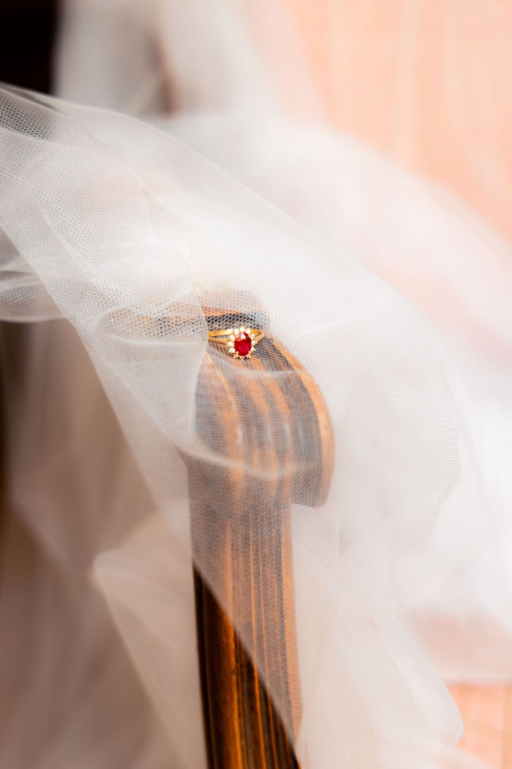 Gold ring with red gemstone resting on wooden chair draped with white tulle fabric at a wedding.