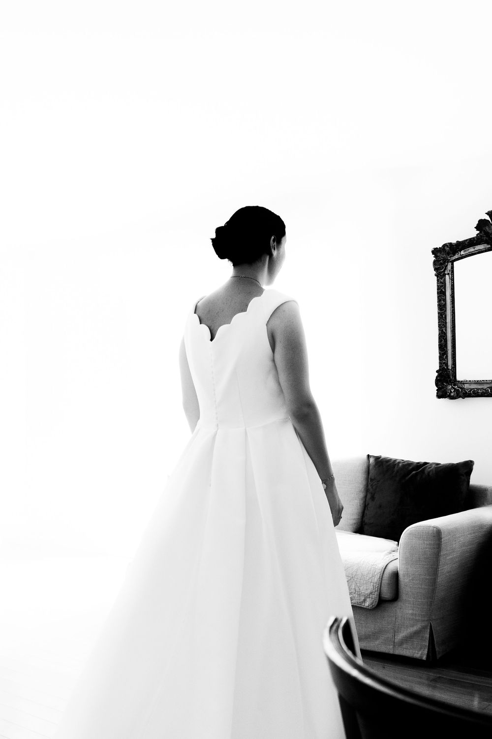 Black and white photo of a bride in a elegant scalloped back wedding gown standing in a softly lit room.