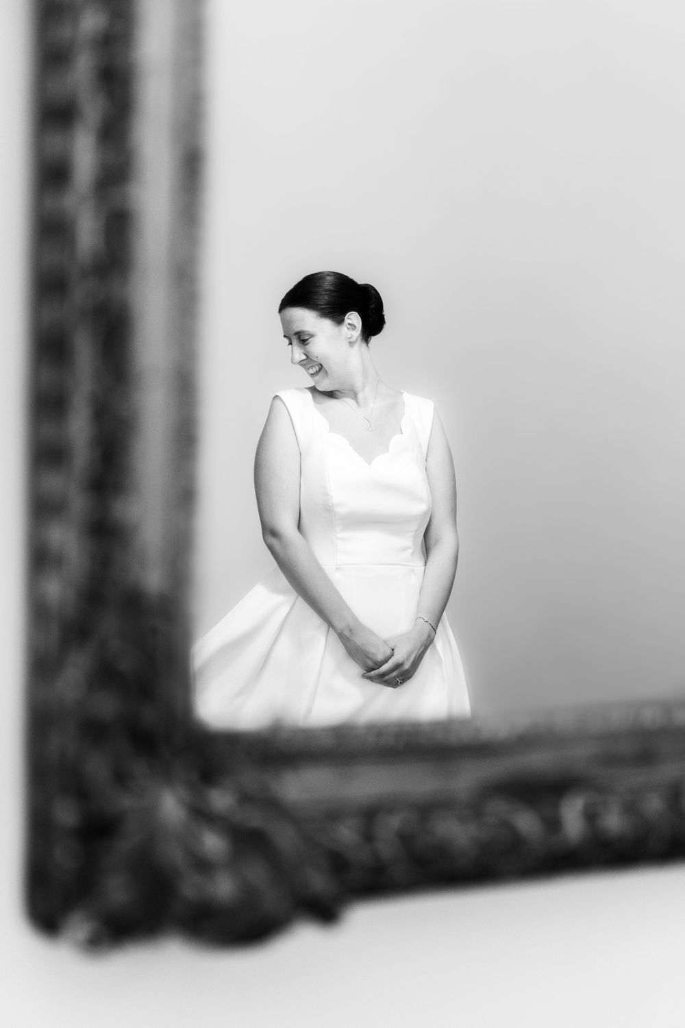 Black and white photo of a smiling bride in a white dress, reflected in an ornate vintage mirror frame.