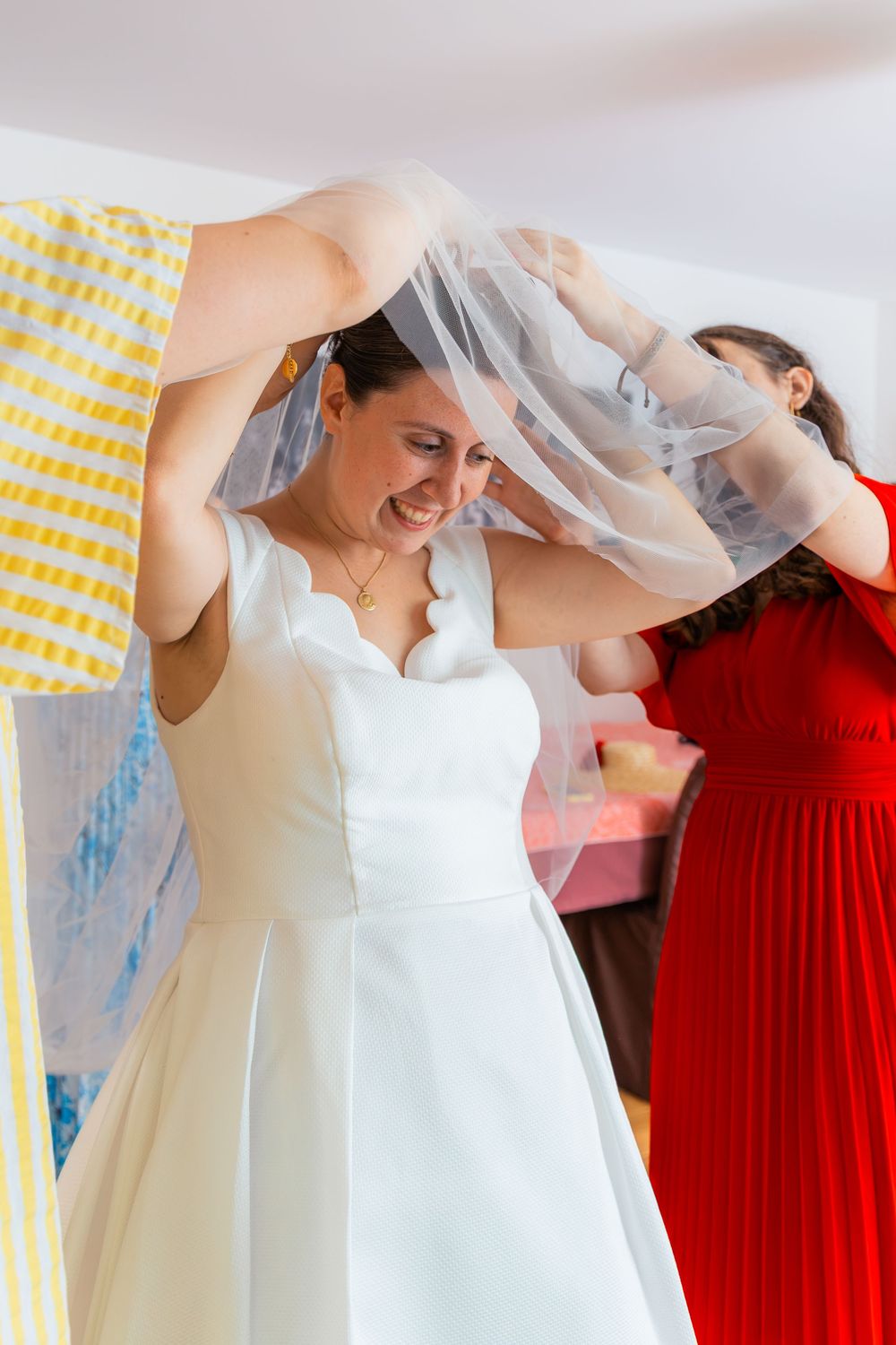 Bride in white gown smiling as bridesmaid in red dress helps arrange her veil during wedding preparations indoors.