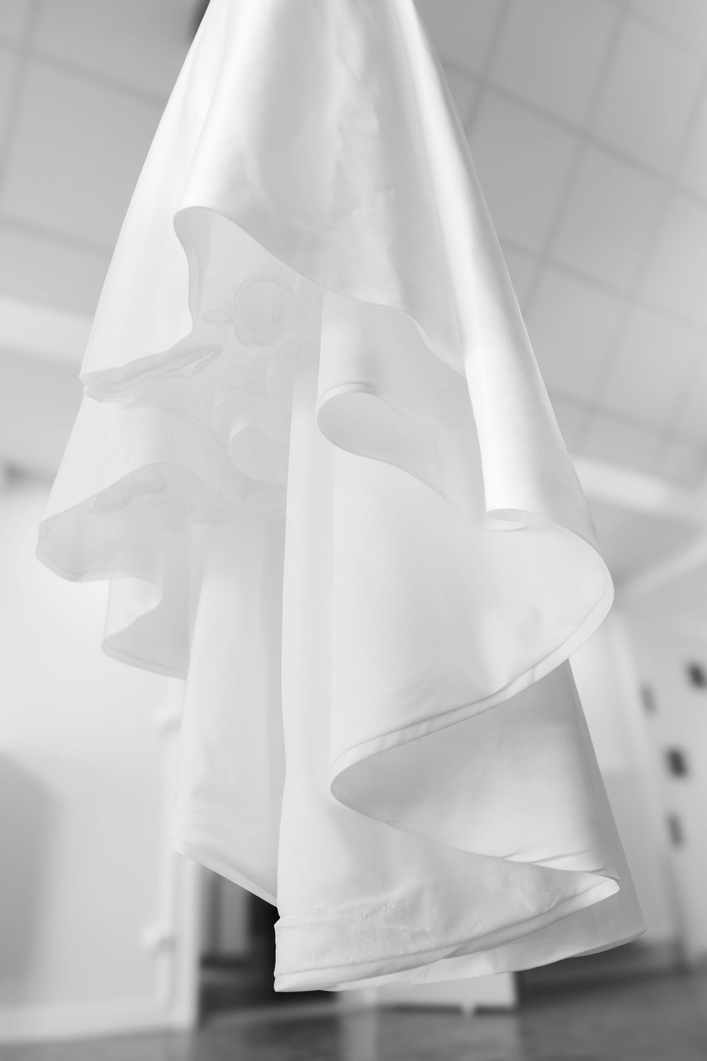 Black and white photo of an elegant white wedding dress with flowing ruffled skirt hanging in a bright room.