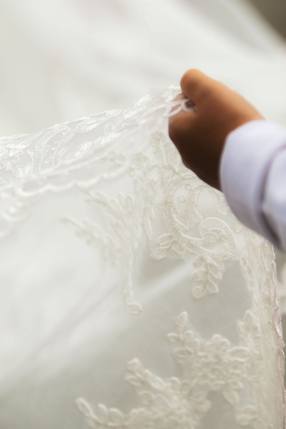 A hand gently holds delicate white lace fabric of a bridal wedding gown with intricate floral embroidery details.