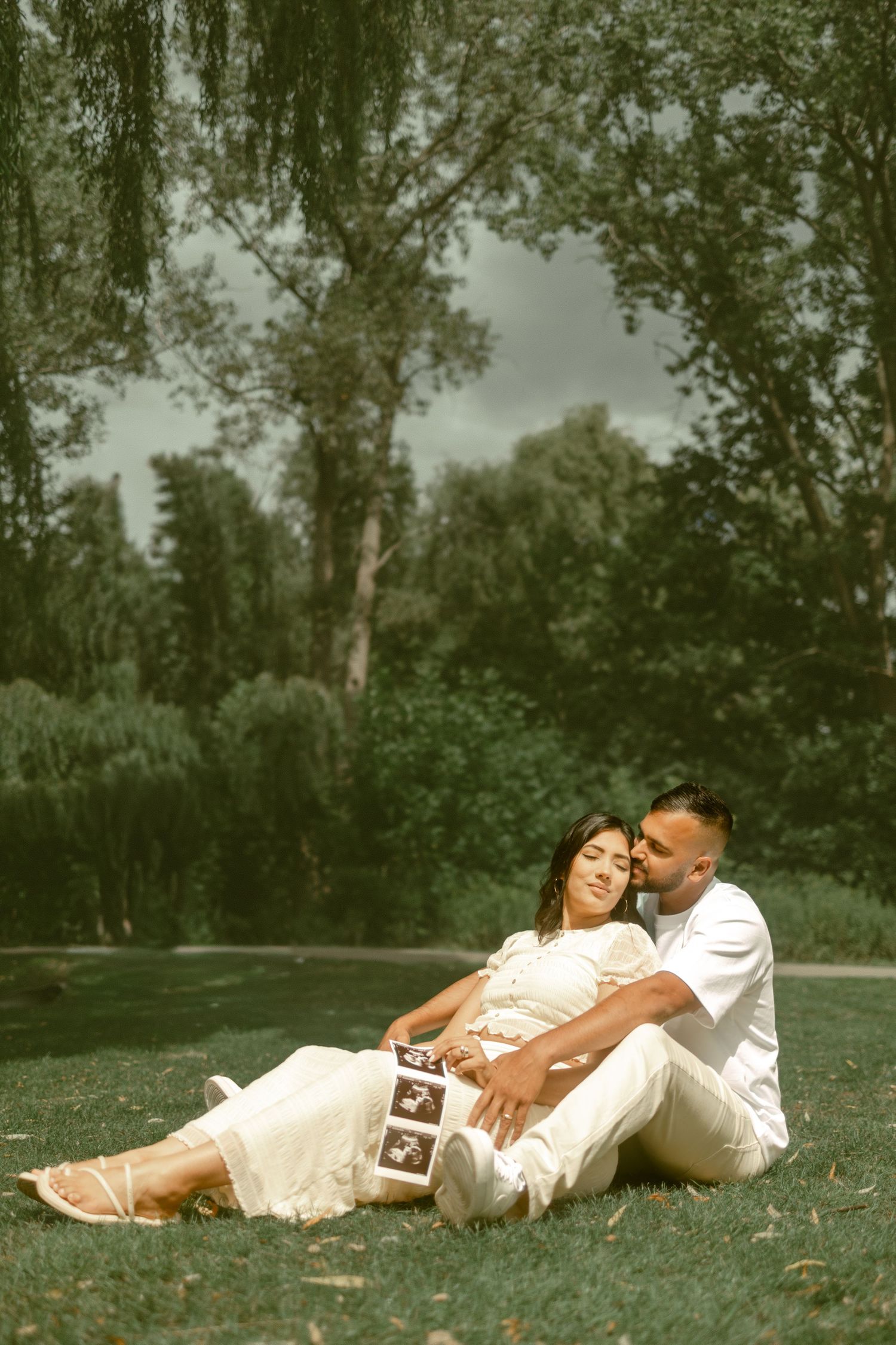 Pregnant couple sitting on grass in park, man kissing woman's cheek, holding ultrasound photos, wearing white outfits.