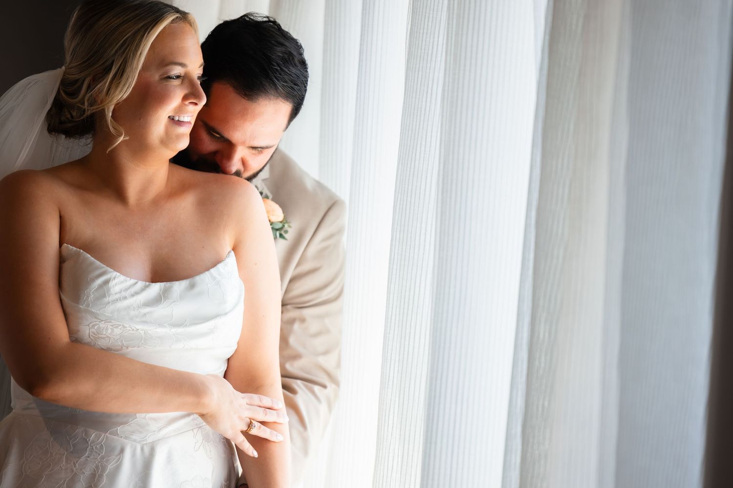 A bride and groom share a tender embrace near a sunlit window on their wedding day.
