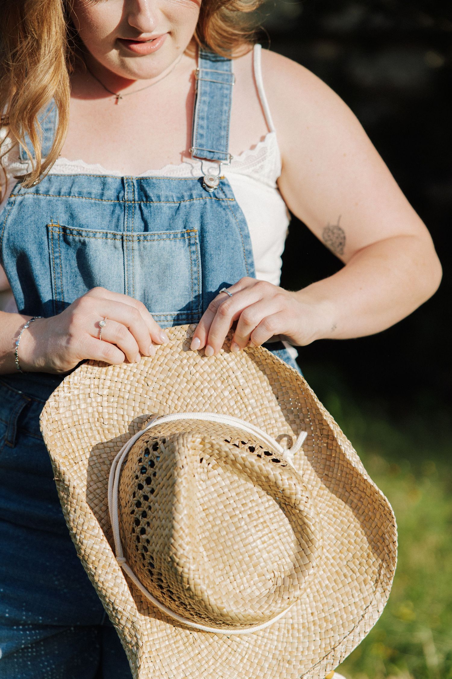 Woman in denim overalls holding a straw cowboy hat outdoors in warm sunlight.