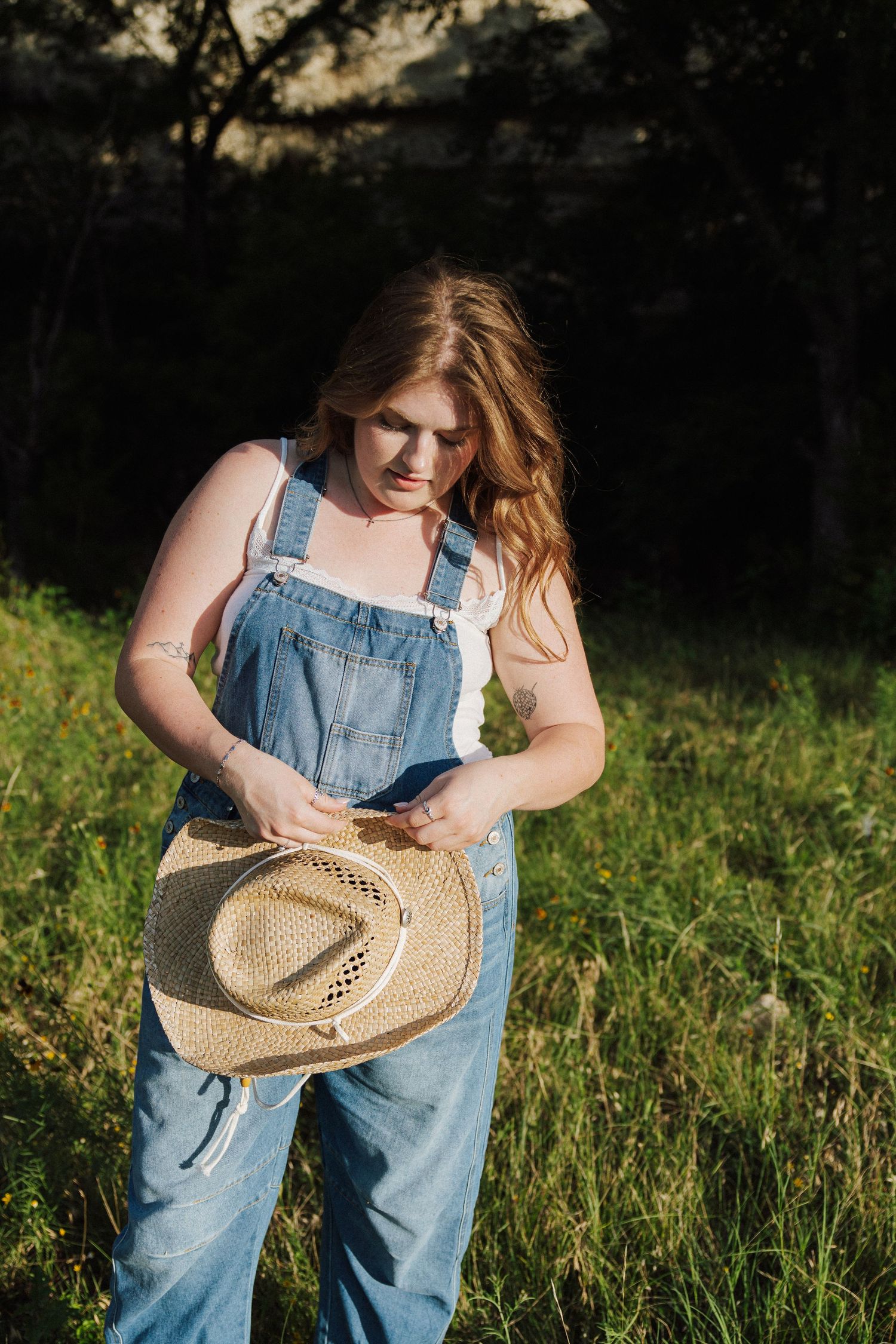 Young woman in denim overalls holding a straw cowboy hat in a sunny green field.