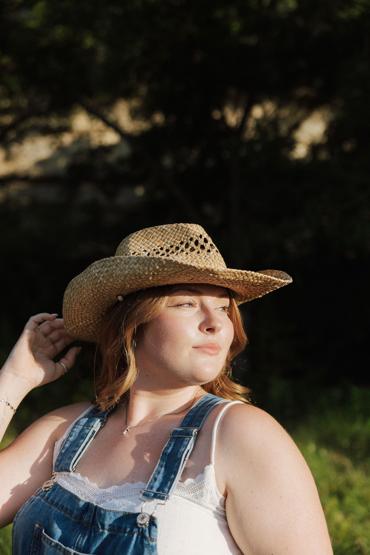 Young woman in straw cowboy hat and denim overalls enjoying golden sunlight outdoors.