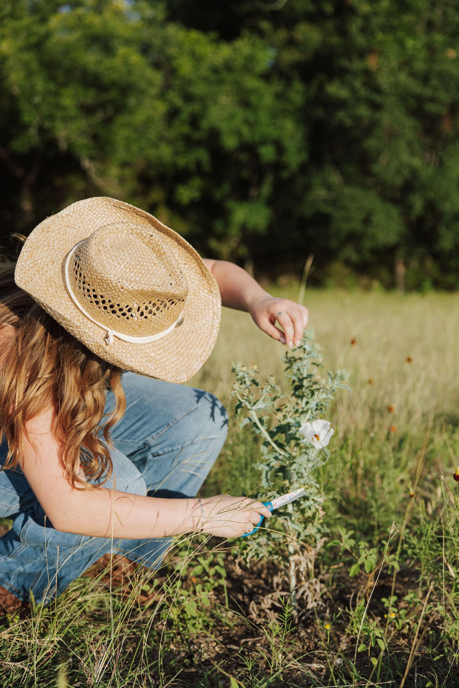 Woman in denim overalls and straw hat picking wildflowers in a sunny meadow.