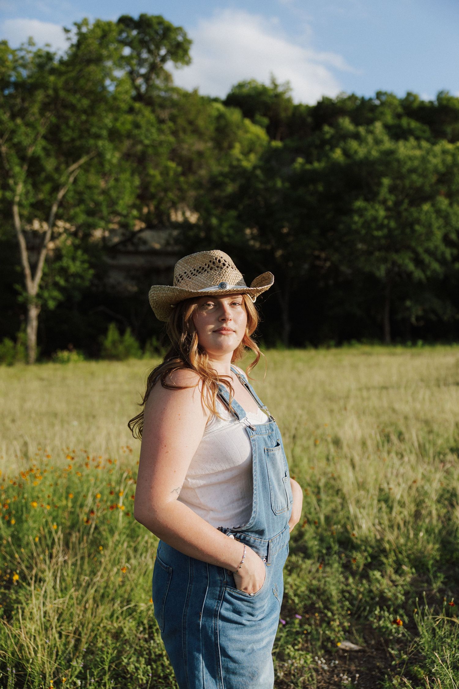 Young woman in denim overalls and straw cowboy hat stands smiling in a sunny green meadow.