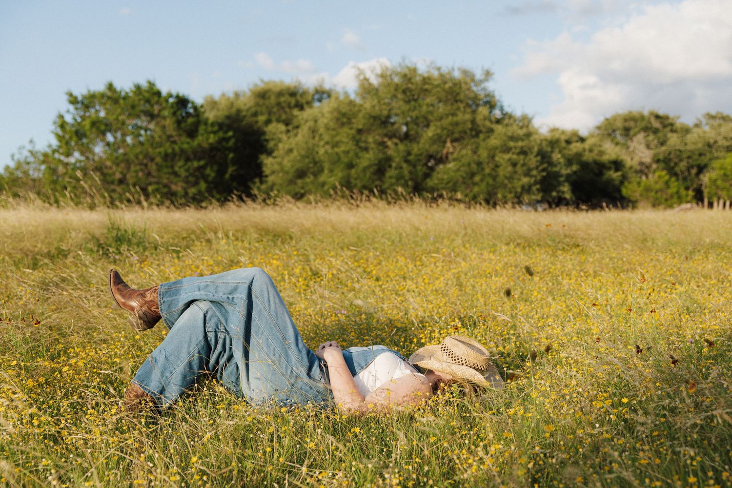 Person lying relaxed in a sunny wildflower meadow wearing jeans, surrounded by lush green grass and trees.