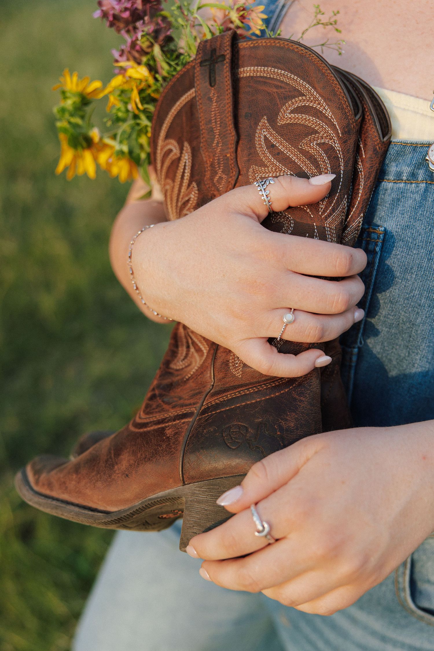 Woman holding brown cowboy boots filled with wildflowers, wearing a diamond ring and delicate bracelet.