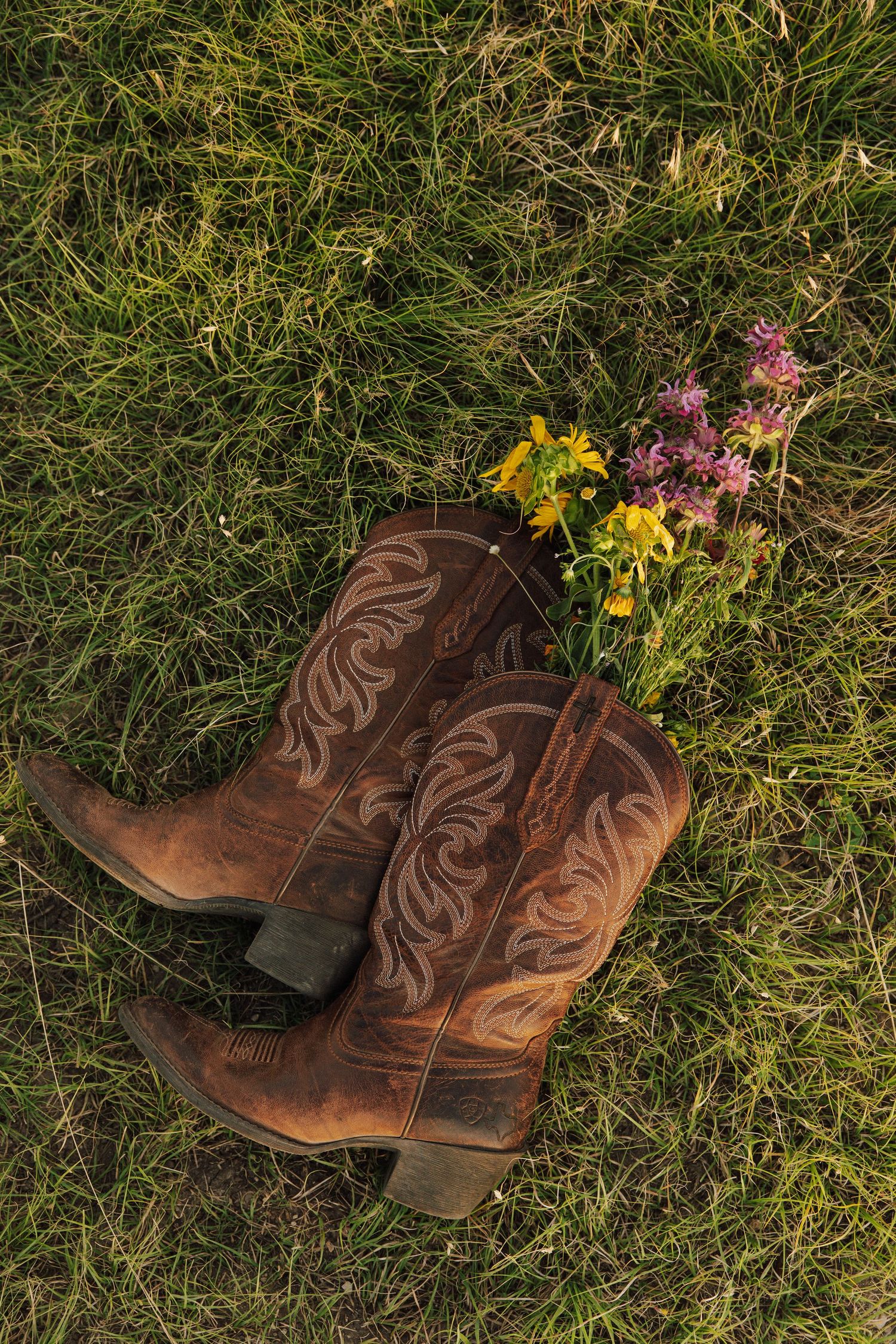 Brown cowboy boots with colorful wildflowers tucked inside resting on green grass.