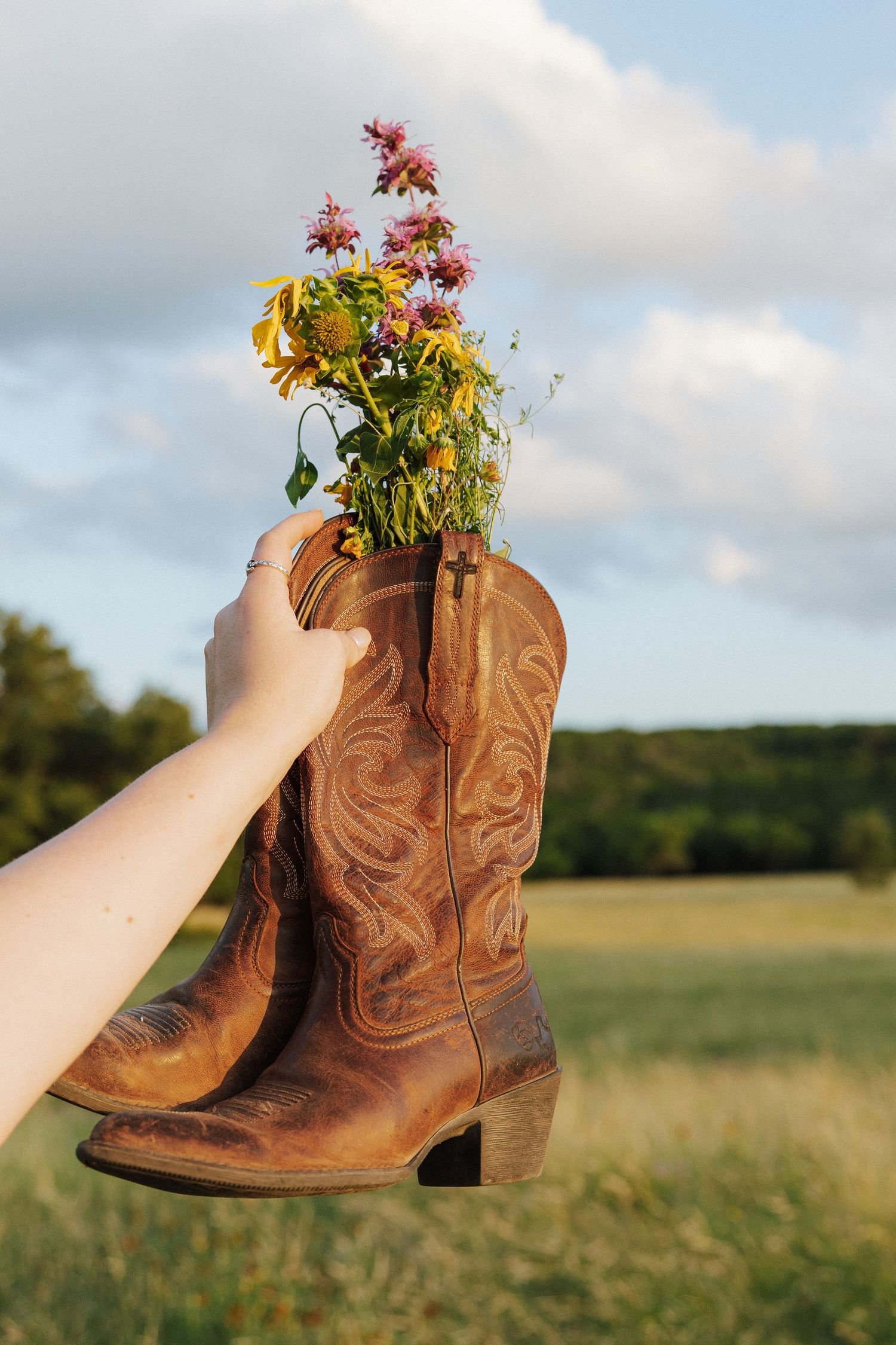 A hand holds brown leather cowboy boots filled with colorful wildflowers against a green field and cloudy sky.