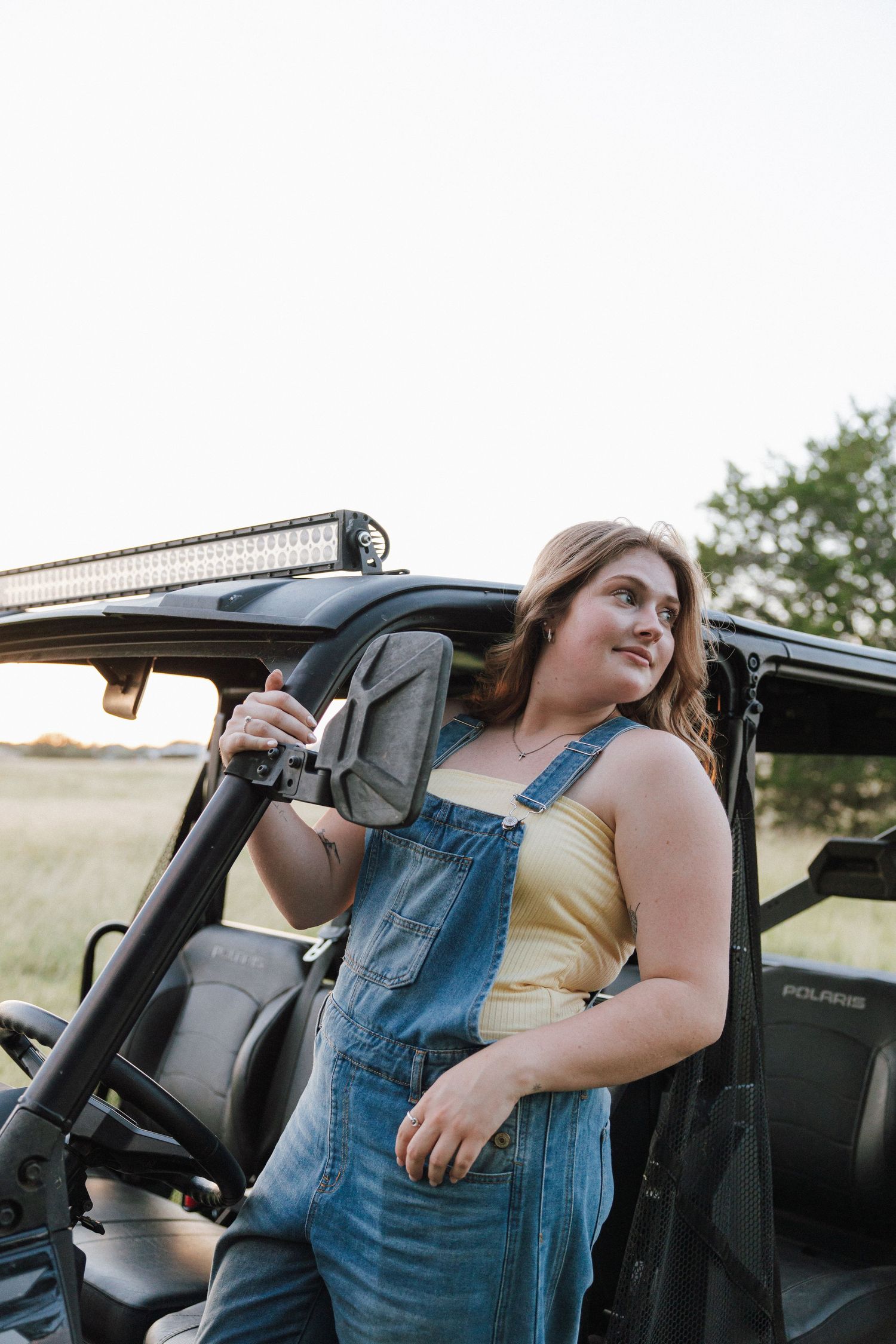 Young woman in denim overalls leaning against a Polaris UTV in an open field at sunset.