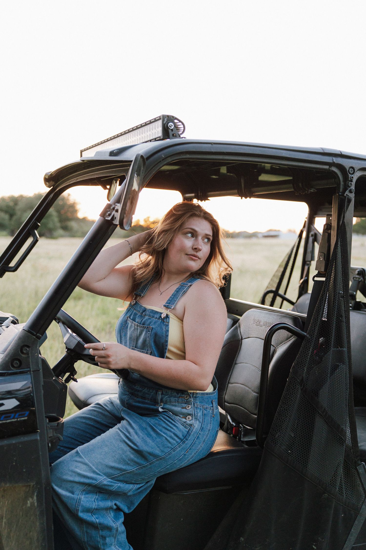 Woman in denim overalls sitting in an off-road UTV vehicle at sunset in a field.