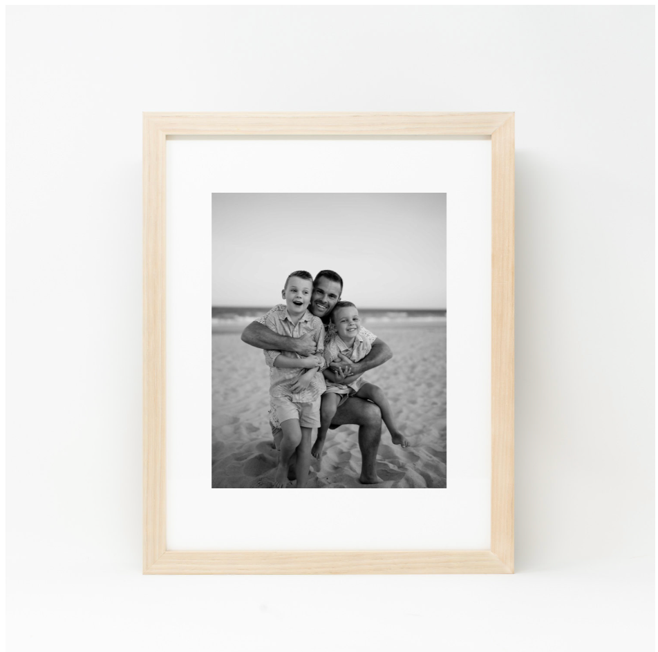 Black and white family portrait in natural wood frame, father holding two young children on a sandy beach.