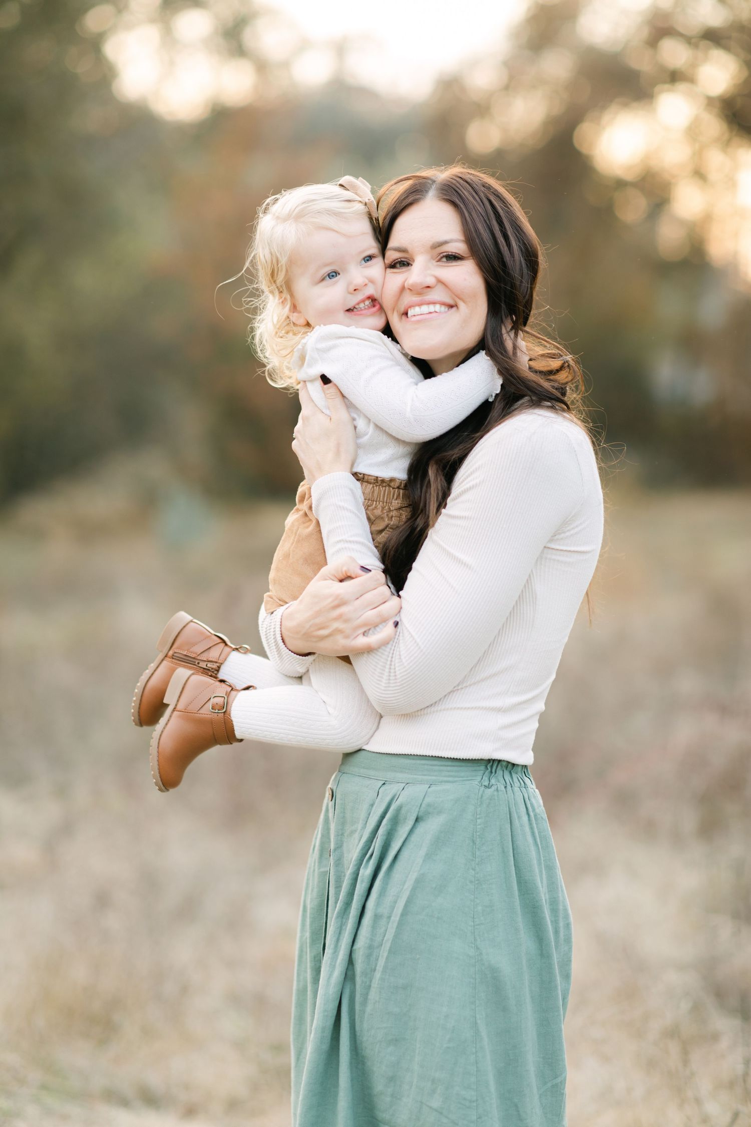 A smiling mother holds her happy toddler outdoors in a sunlit field, both dressed in warm autumn tones.
