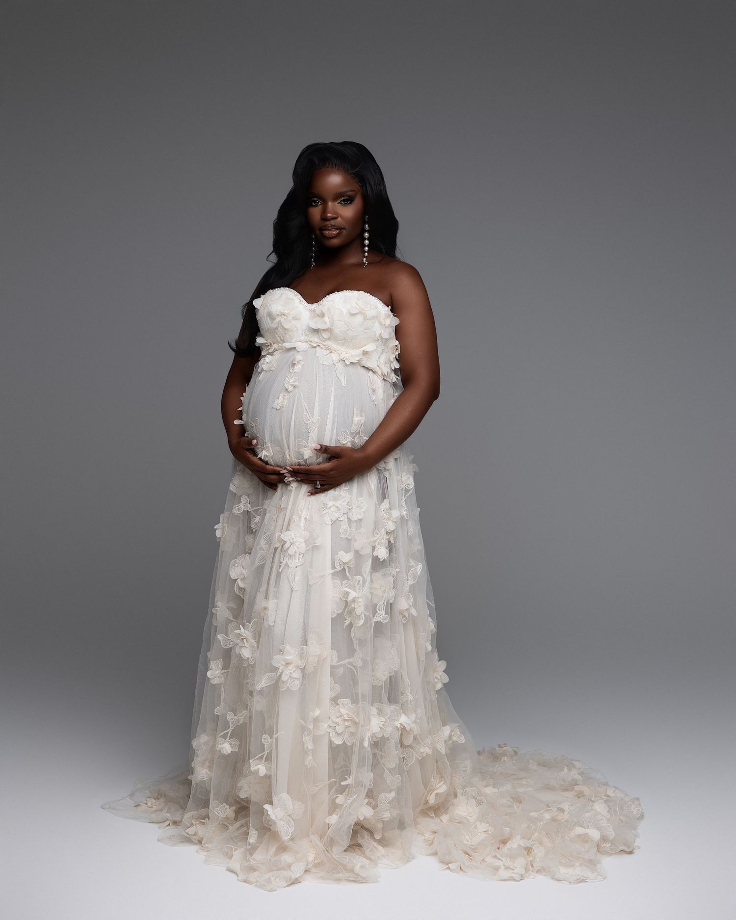 Pregnant woman in elegant white floral tulle strapless gown posing gracefully in a professional maternity photo shoot.