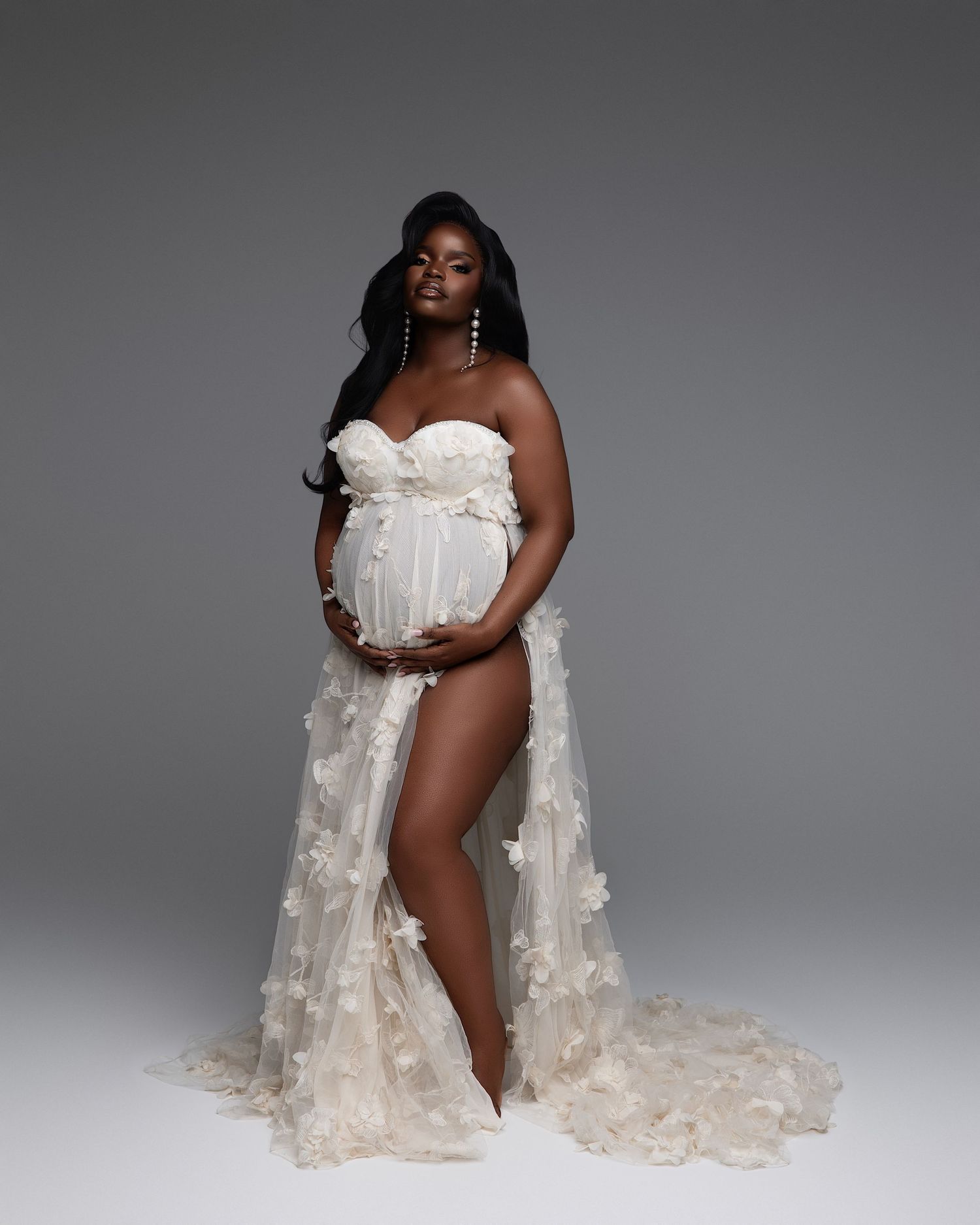 Pregnant woman in elegant white strapless floral gown with sheer train, posing gracefully in a professional maternity photo shoot.