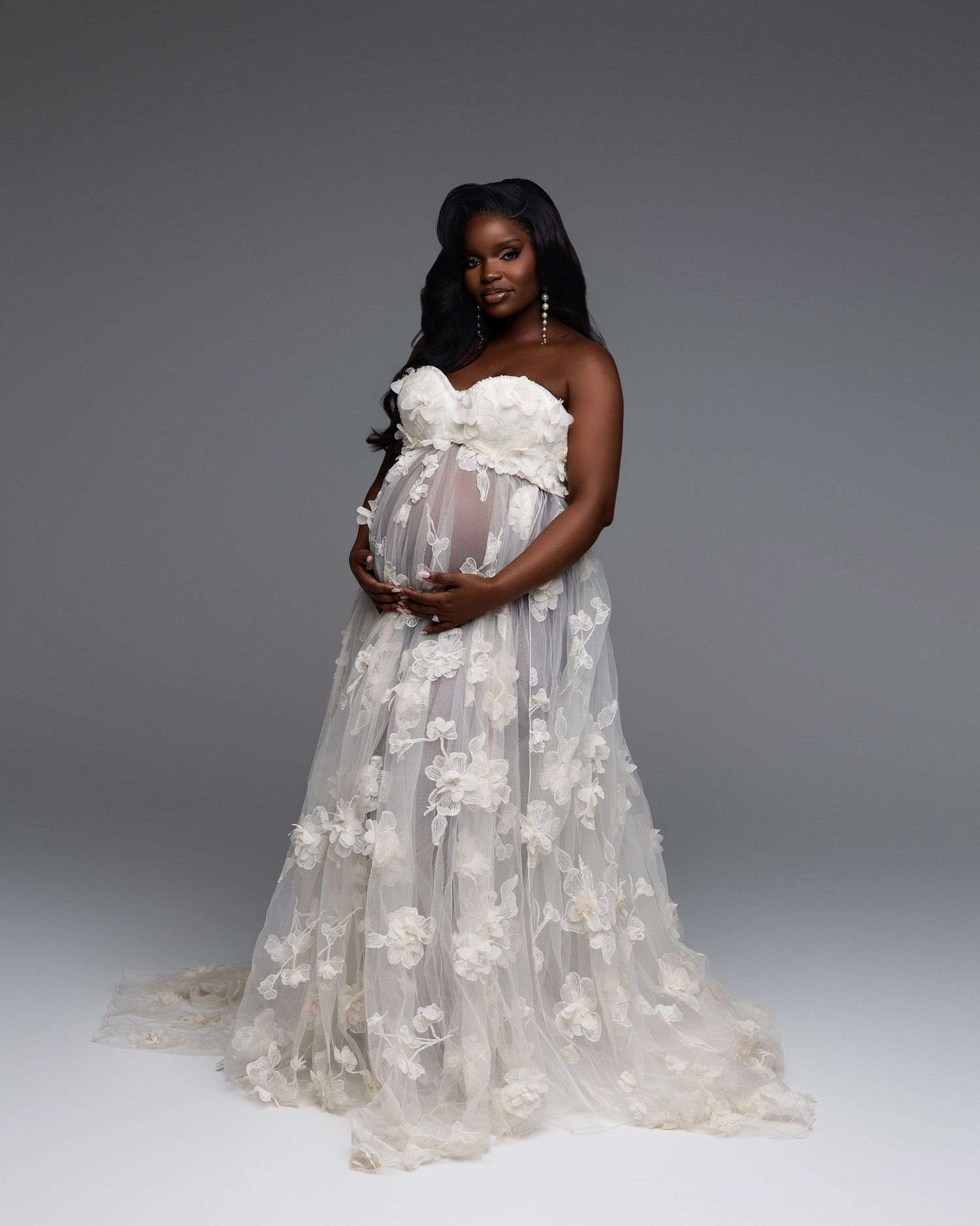 Pregnant woman in white floral tulle maternity gown posing gracefully in a studio, cradling her baby bump.