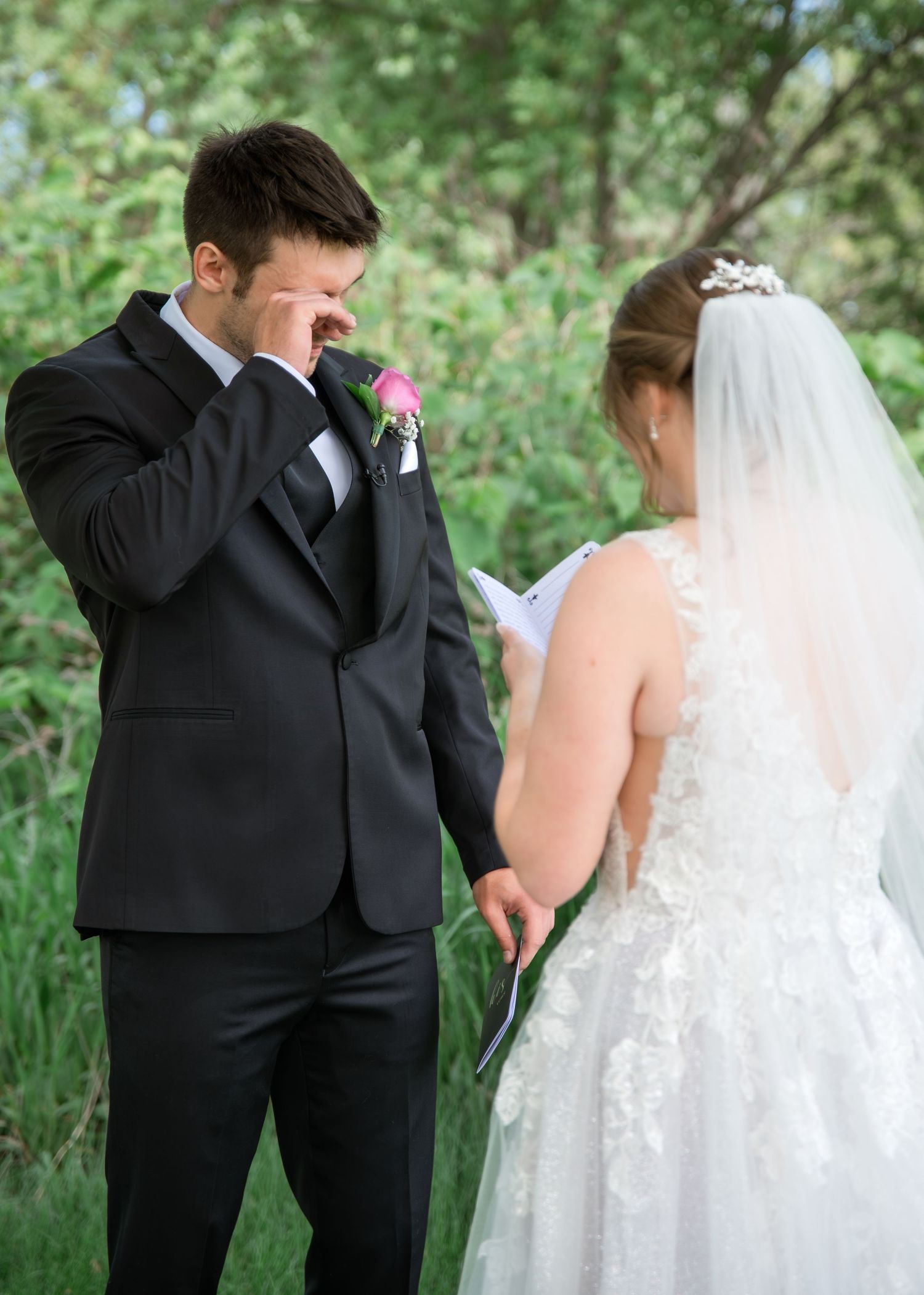 Bride in lace gown with cathedral veil embraced by groom in black suit in lush green garden setting.