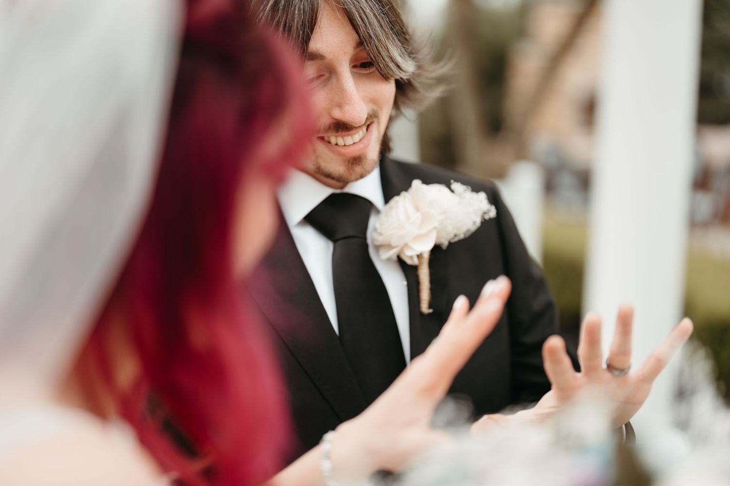 Smiling groom in black suit with white boutonniere looks at red-haired bride during outdoor wedding ceremony.