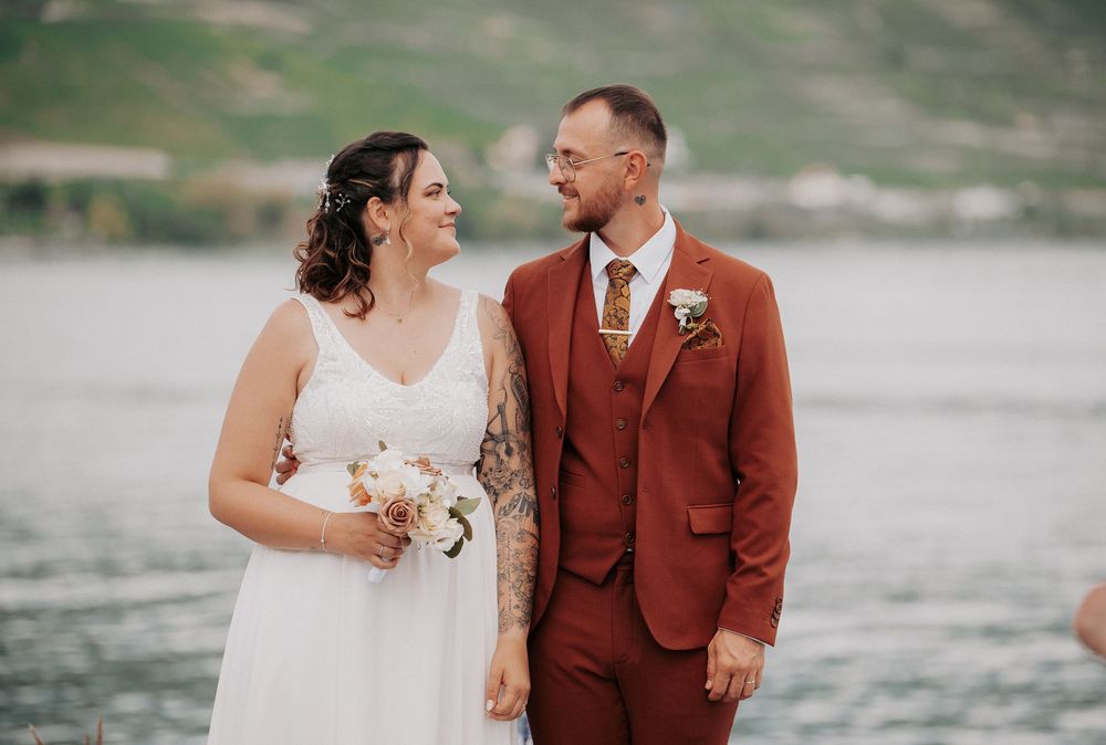 A couple in wedding attire poses by a scenic waterfront with mountains in the background.