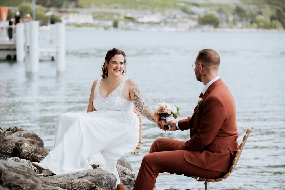 A couple in wedding attire sits together on rocks by a serene lake.