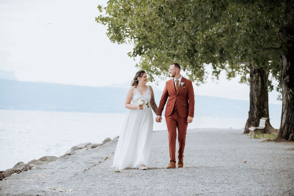 A couple walks hand in hand along a tree-lined waterfront path.