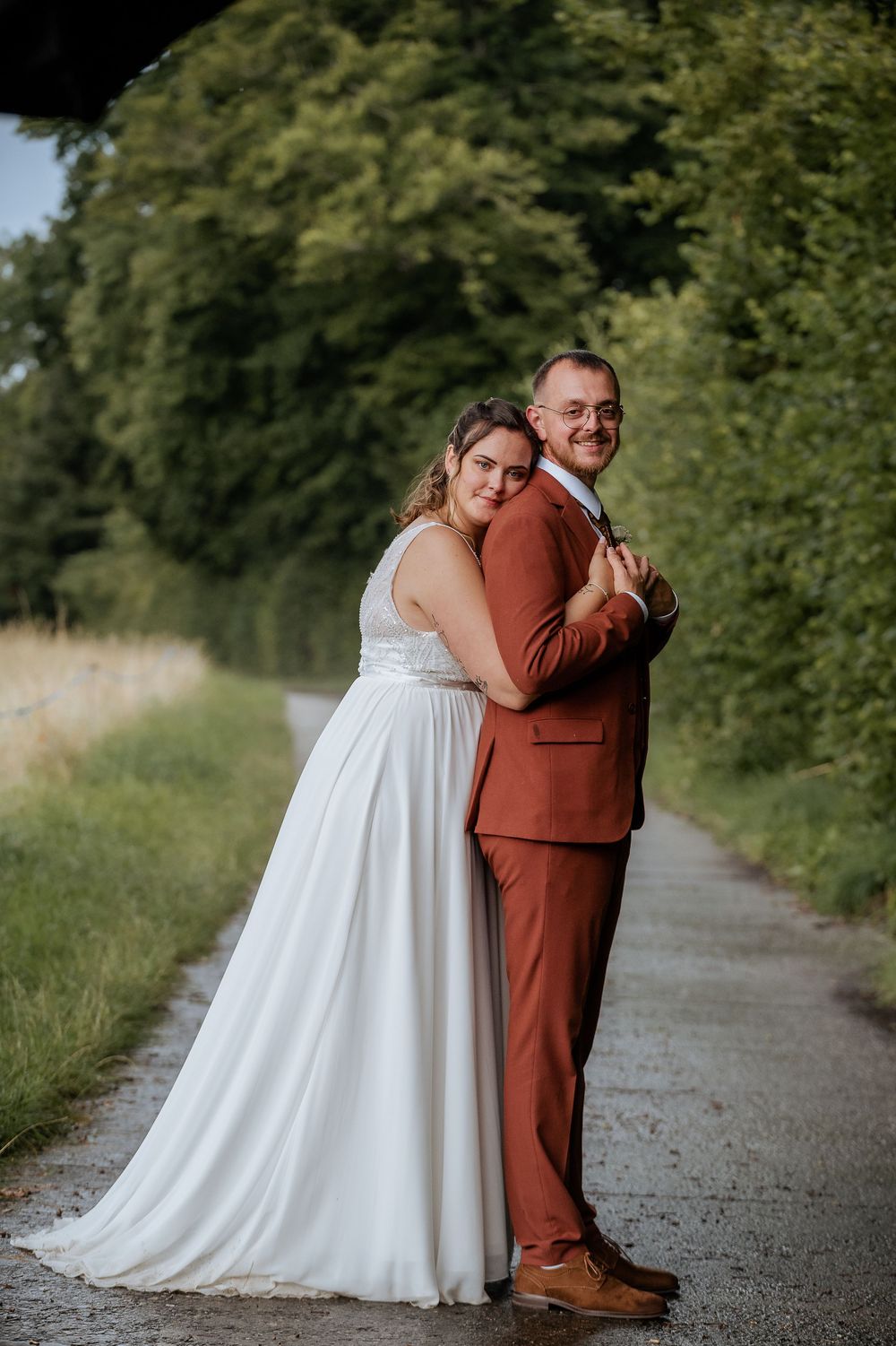 A newlywed couple poses back-to-back on a tree-lined path, bride in flowing white dress and groom in rust suit.