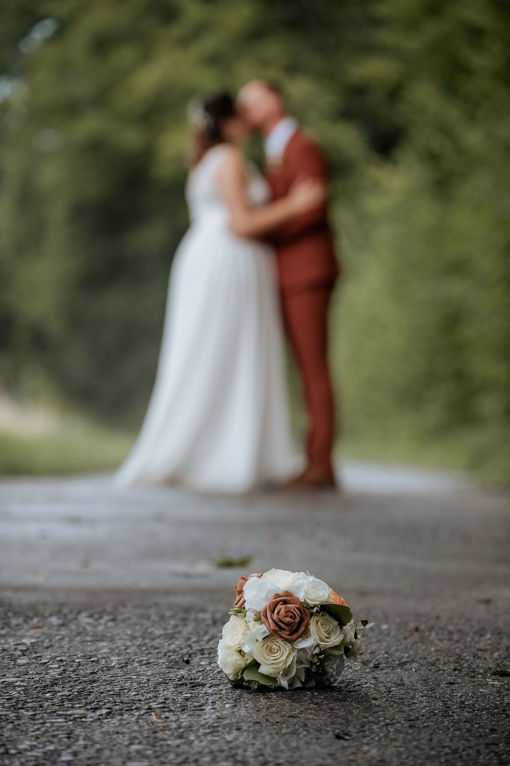 Wedding bouquet lies on gravel path with couple blurred in background.