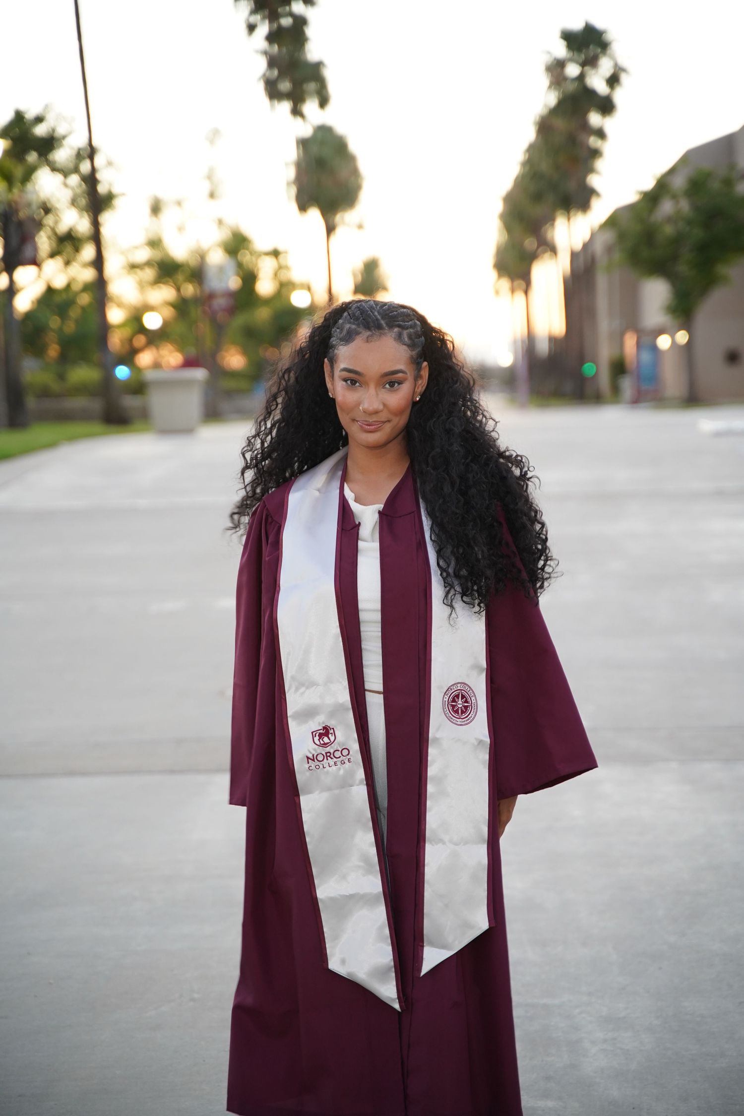 Young Black woman in maroon graduation cap and gown with honor stole stands on palm-lined campus.