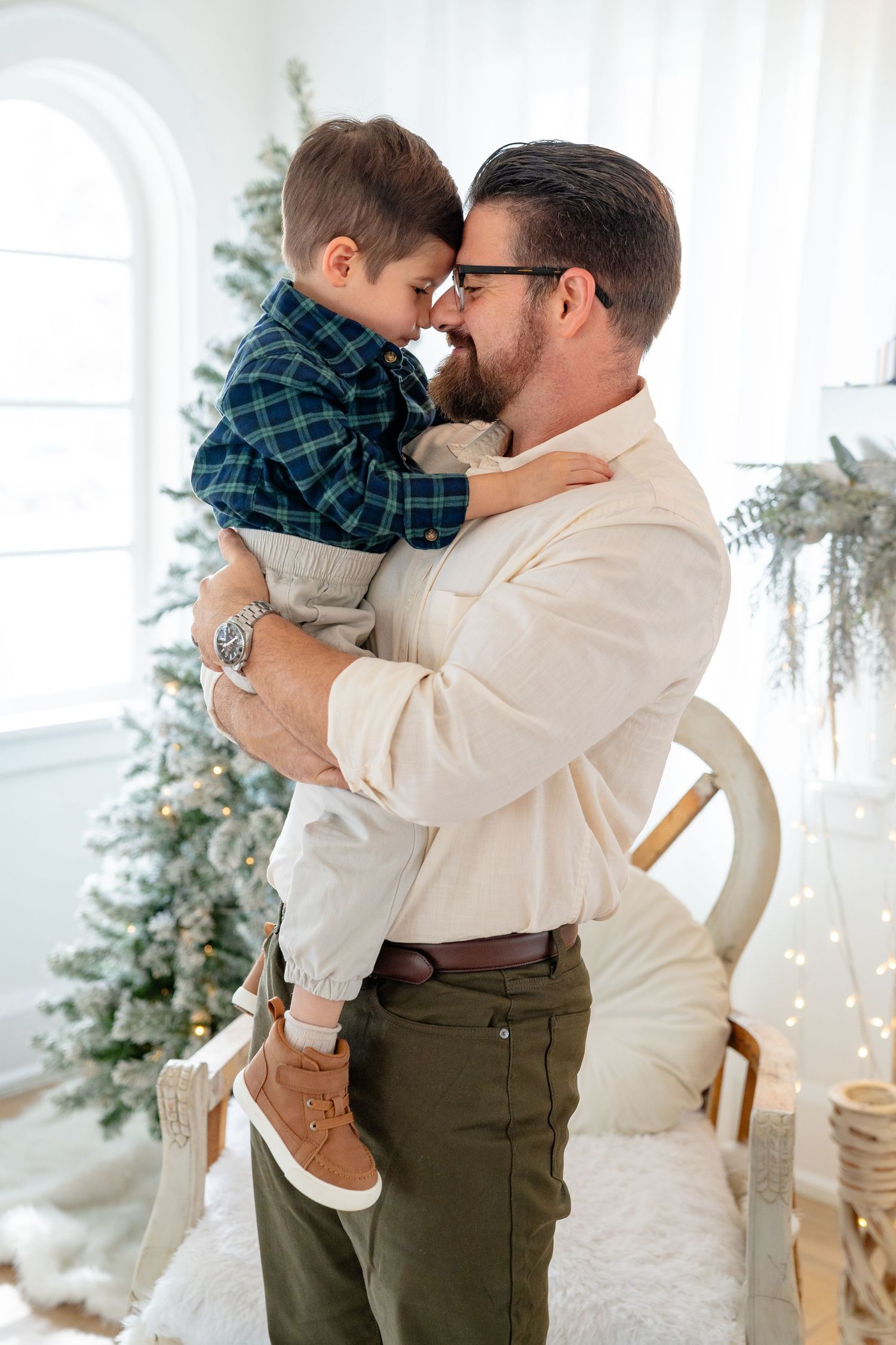 Father holding young son nose-to-nose near decorated Christmas tree in bright white holiday room.