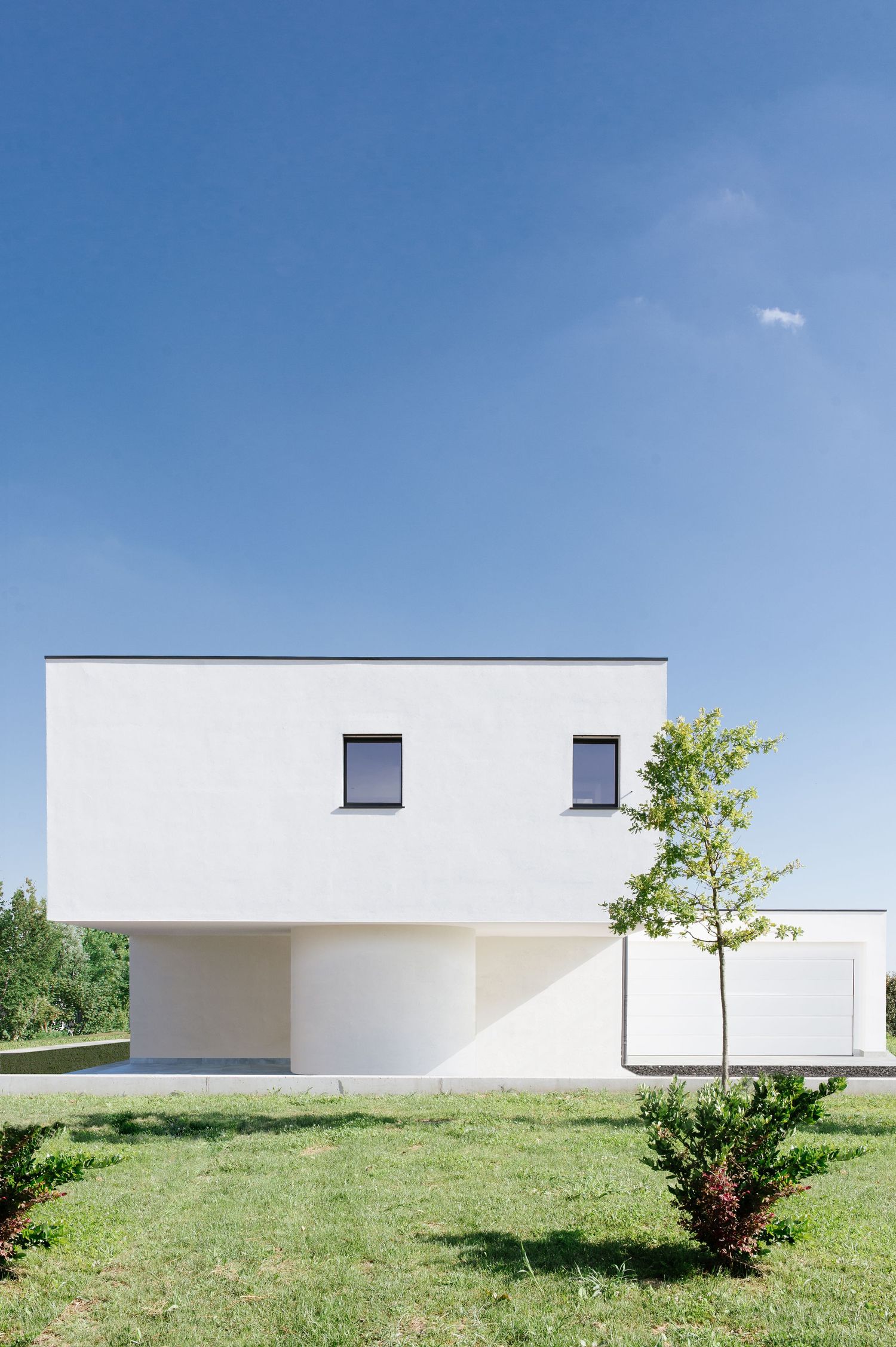 Modern minimalist white cubic house with flat roof, two windows, and small trees on green lawn under clear blue sky.