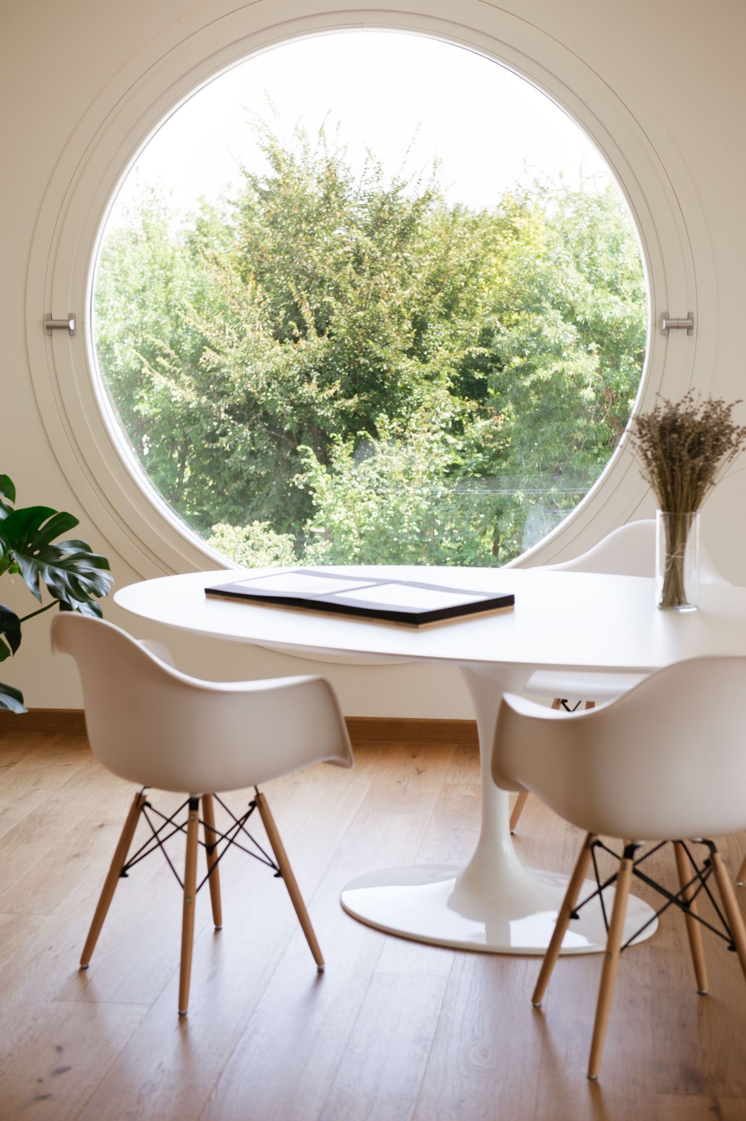 White tulip table and Eames chairs in a minimalist dining nook with a large round window overlooking lush green trees.