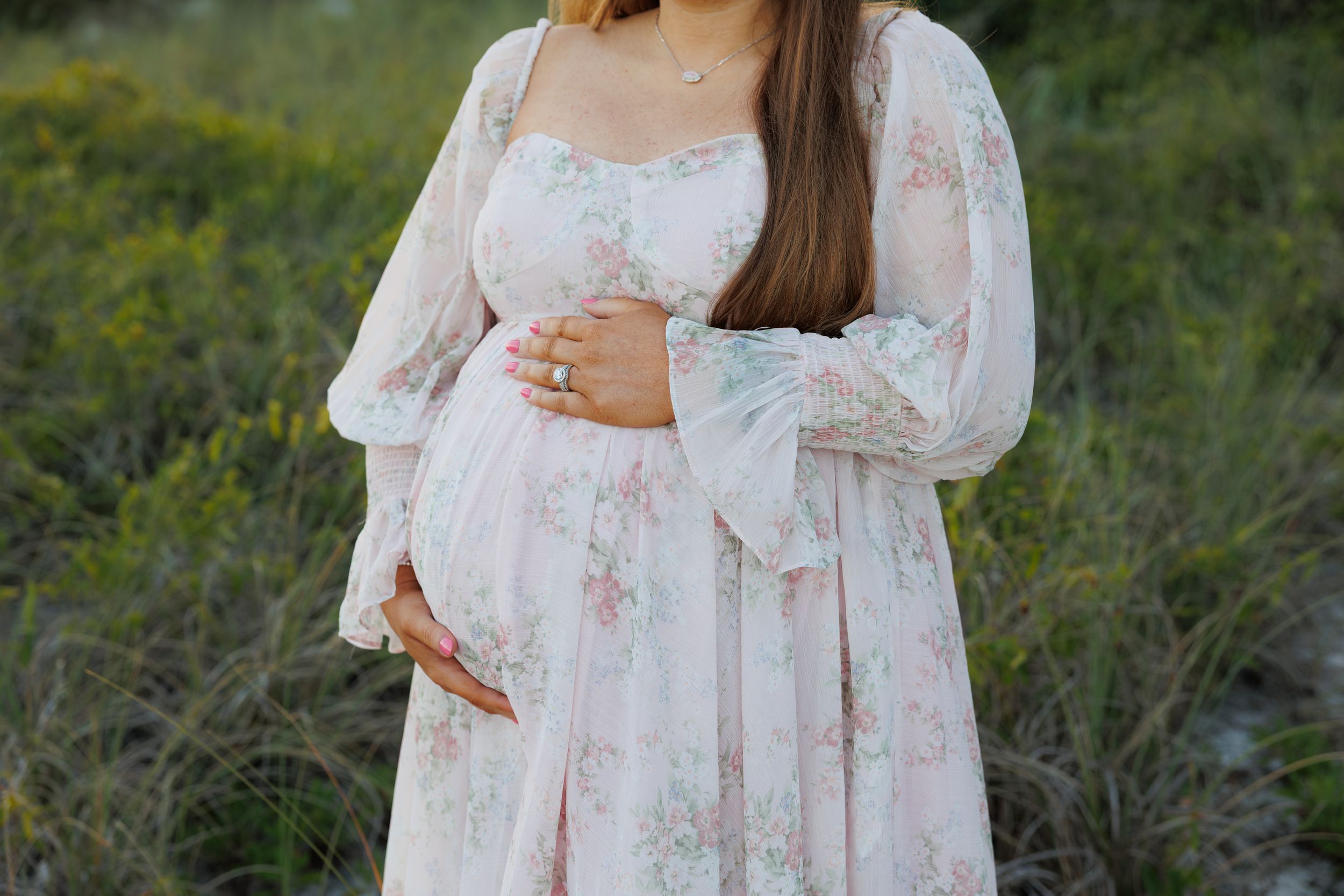 Pregnant woman in floral pink maternity dress holding baby bump outdoors in green field during maternity photo session.