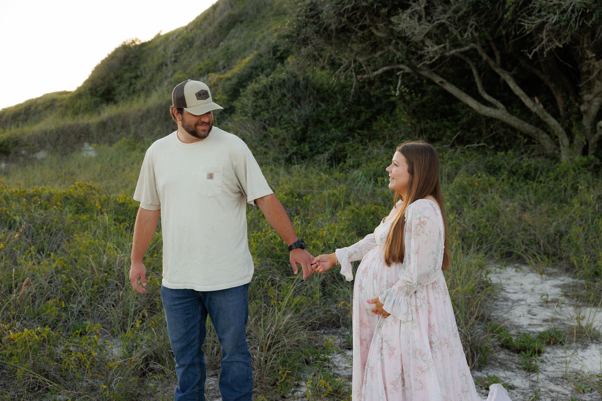 Couple holding hands outdoors near sand dunes and greenery, woman in pink dress appears pregnant.